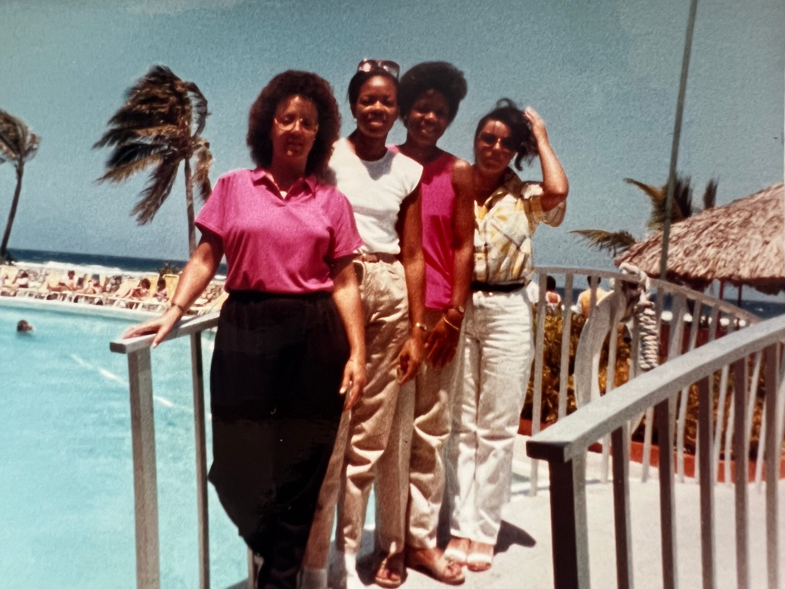 Four women pose together on a sunny beach boardwalk with palm trees and the ocean in the background.