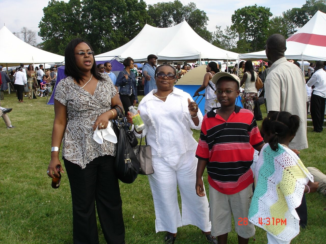 Group of people at an outdoor festival with white tents in the background; woman in patterned top left holding a drink and bag, other woman in white, and two kids in foreground.