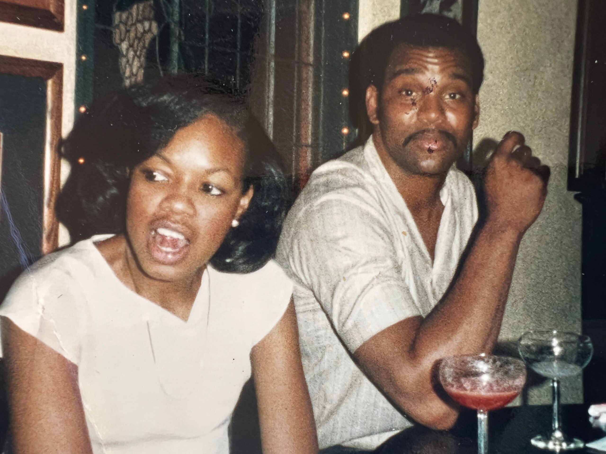 Two friends at a dinner table, the woman laughing with mouth open while the man raises a glass of red beverage in a toast.