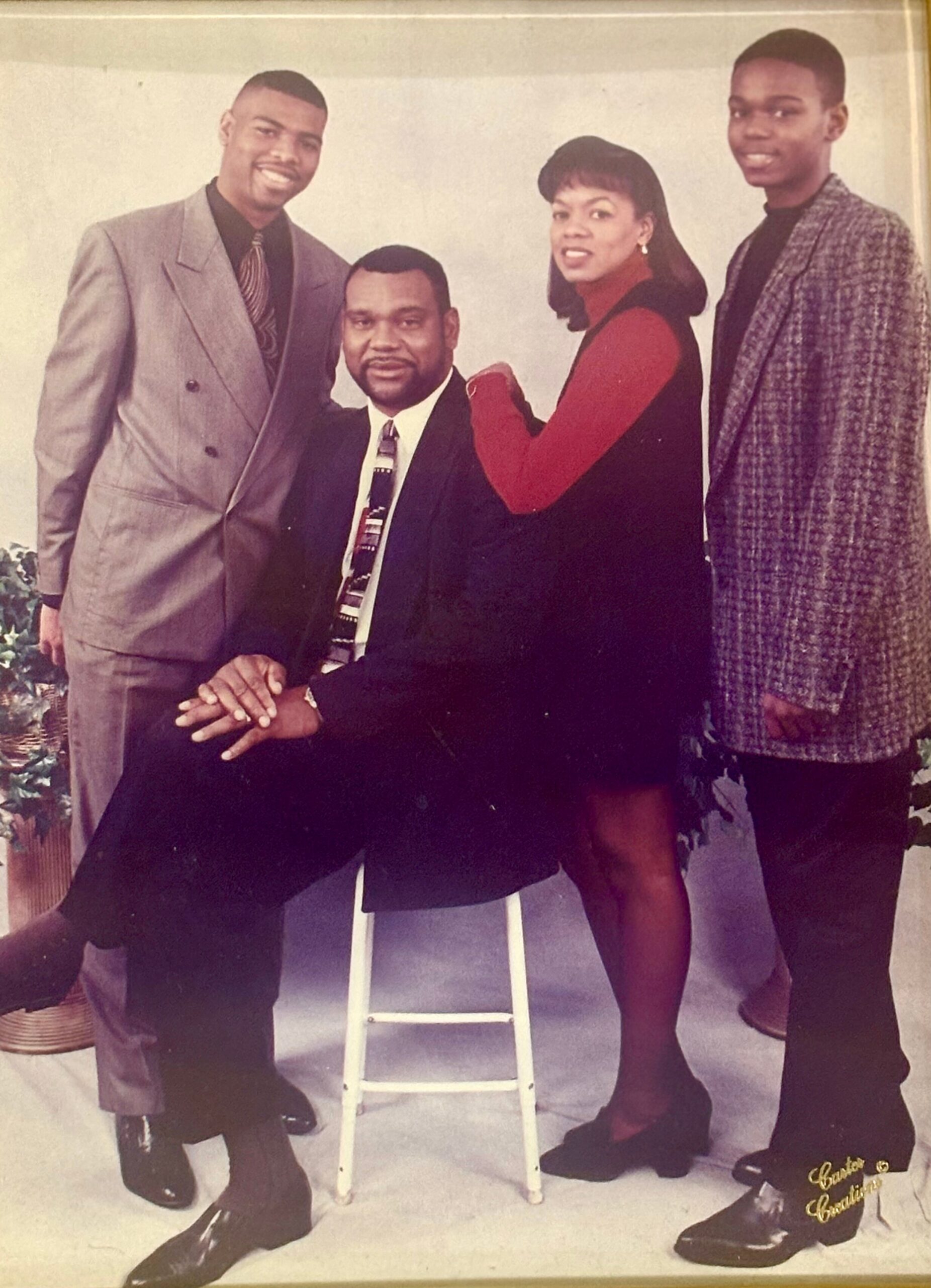 Family portrait with four people: a seated man in a suit, a woman in red and black vest beside him, and two men standing on either side in vintage attire.