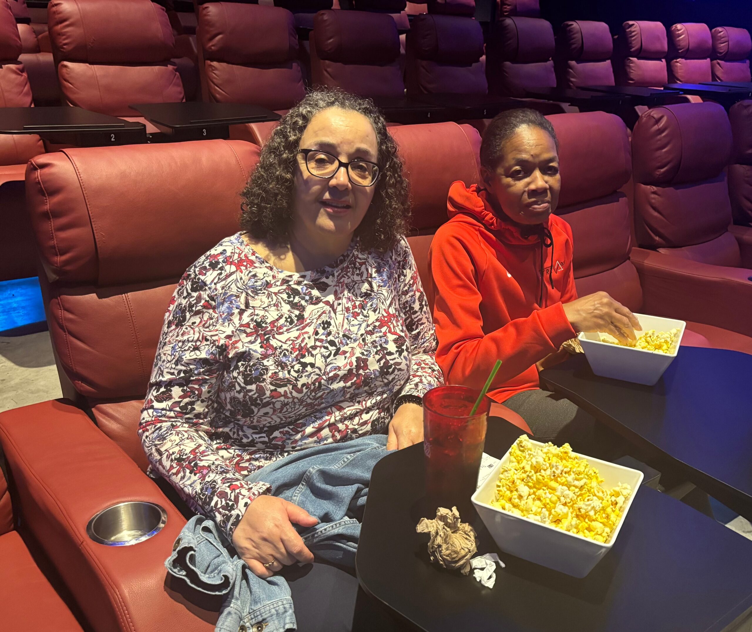 Two people sit in a red theater, sharing popcorn on small tables with drinks nearby, under purple lighting.