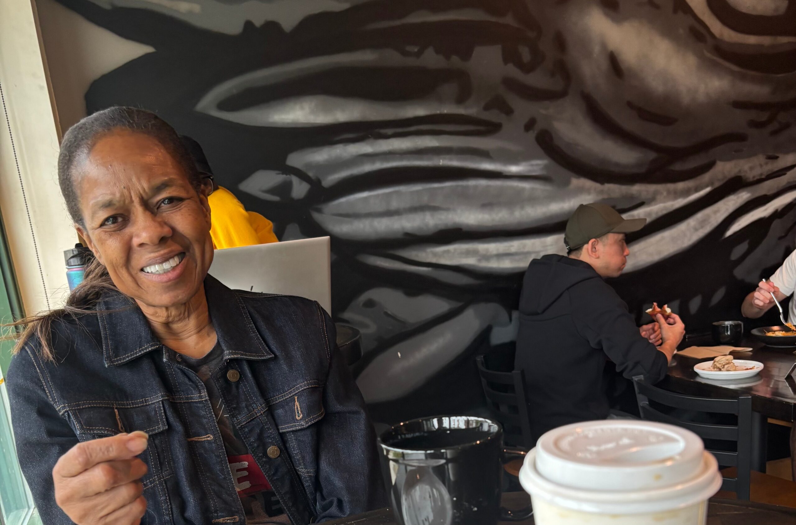 Smiling woman in a denim jacket seated at a table in a cafe with a black-and-white mural behind her.