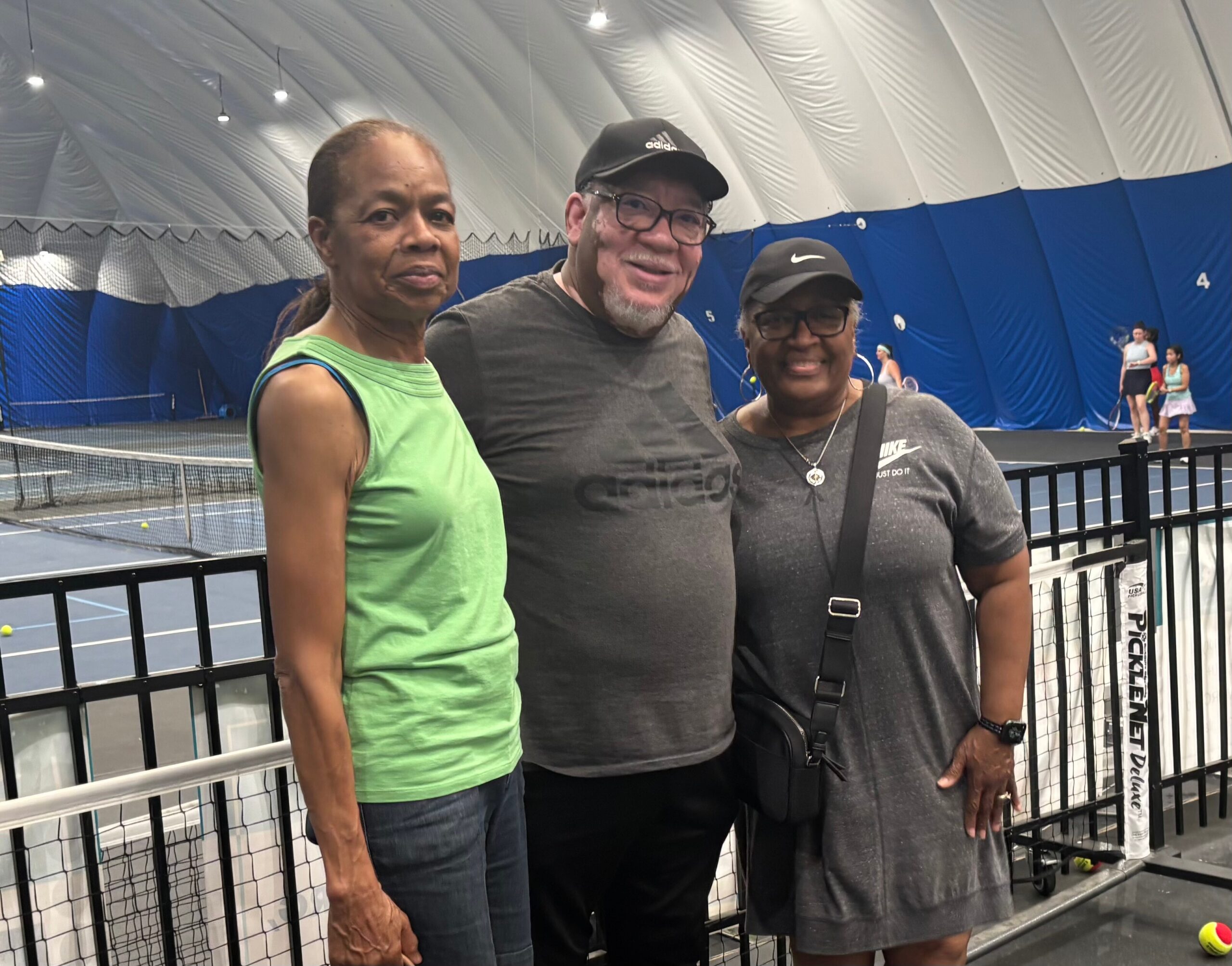Three adults posing together at an indoor tennis facility, smiling with a tennis court and net in the background.