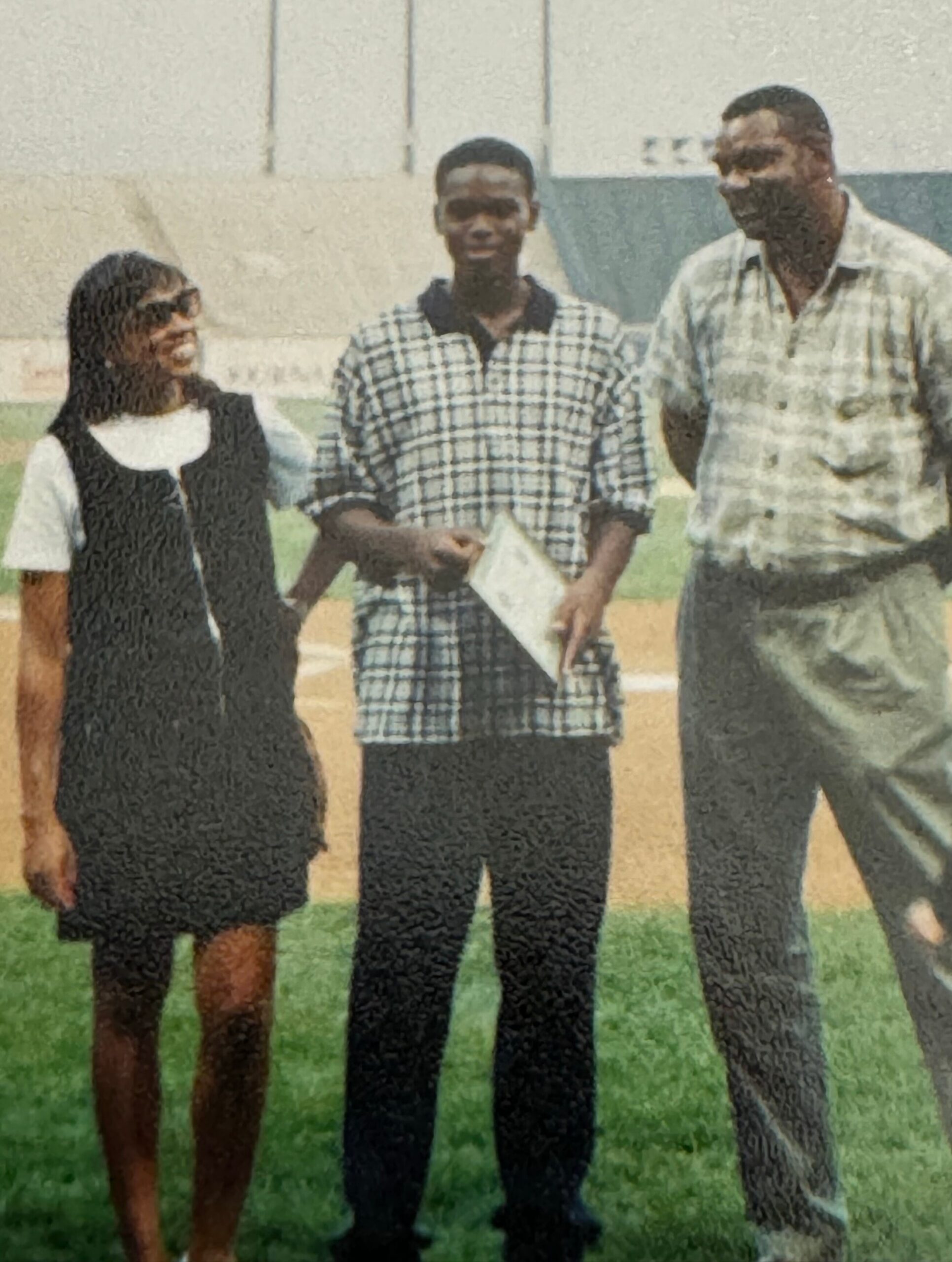 Three people stand side by side on a grassy field, posing for a photo with a building in the background.