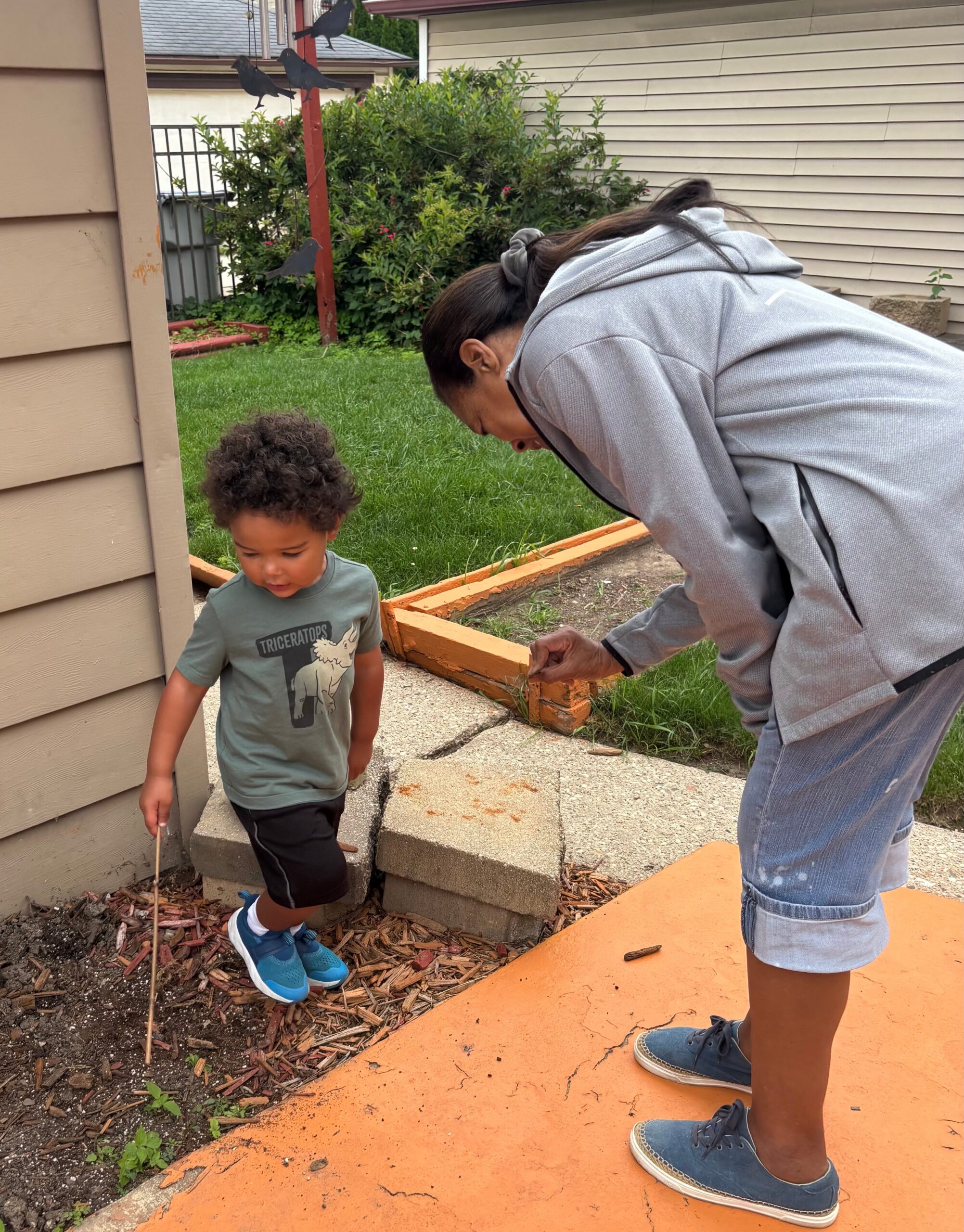 Woman bending over beside a young child, gardening at a raised bed with orange edging outside a house.