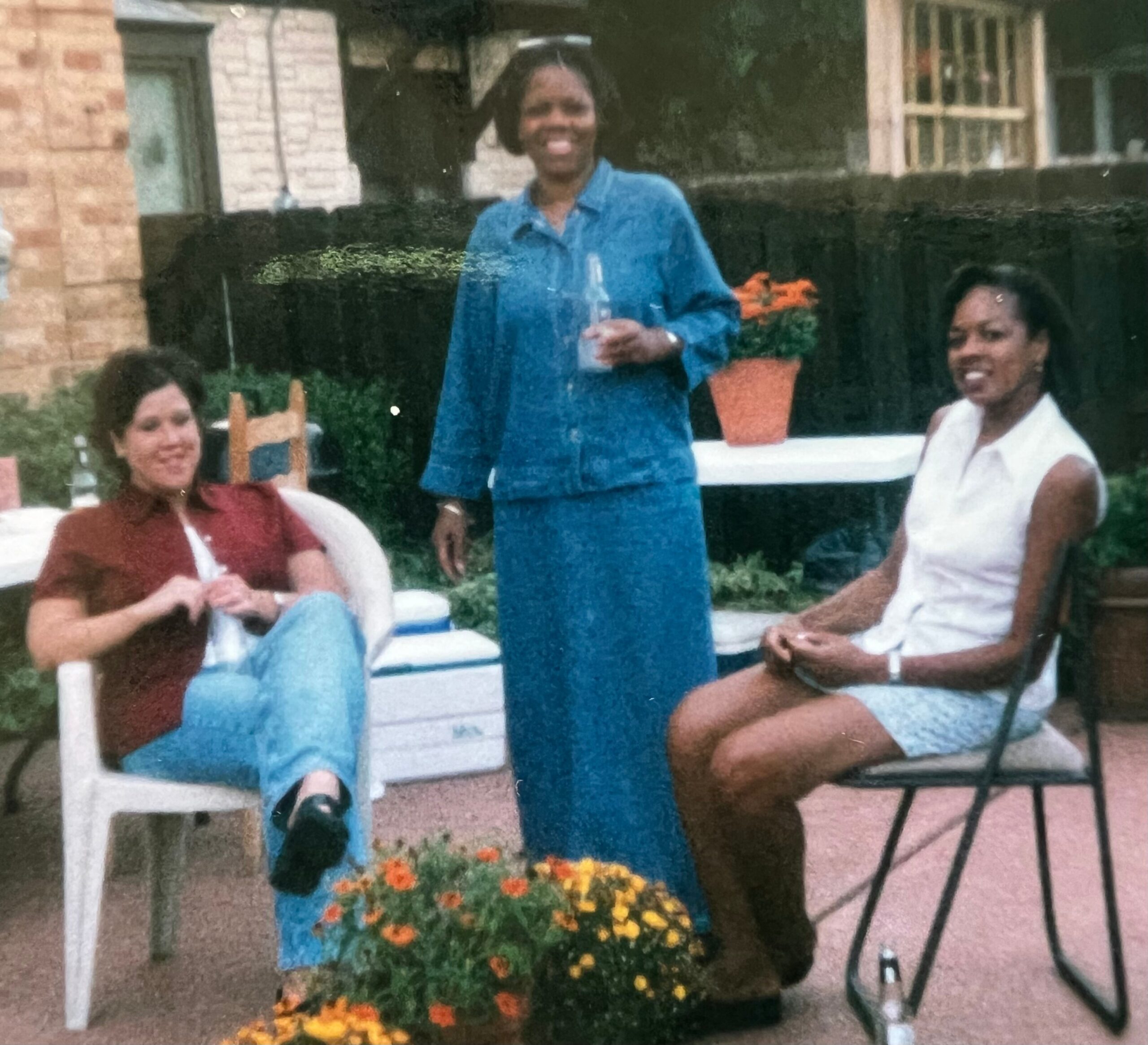 Group of people in a backyard garden; a woman in a blue dress stands holding a bottle, with others seated nearby and orange flowers in the foreground.