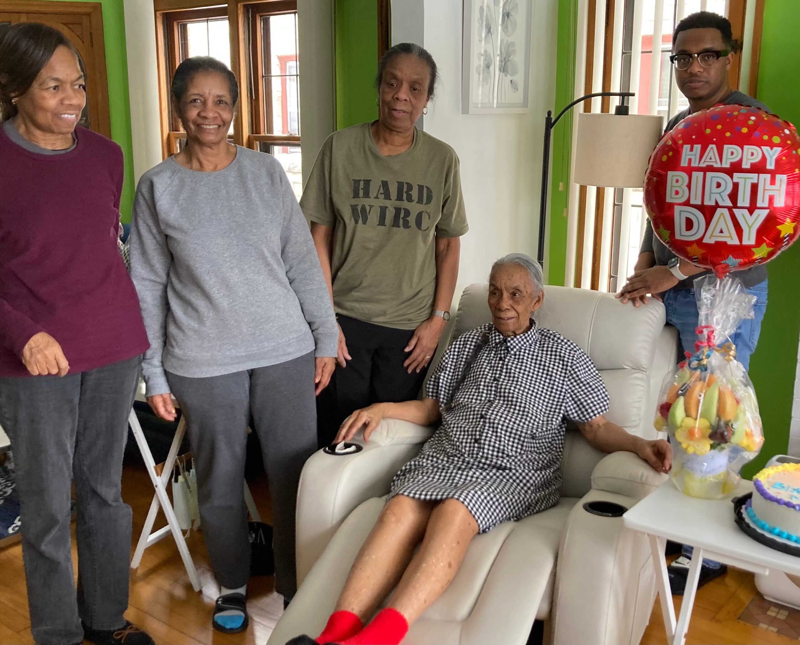 Group of adults posing indoors for a birthday celebration, with a red 'Happy Birthday' balloon visible on the right.