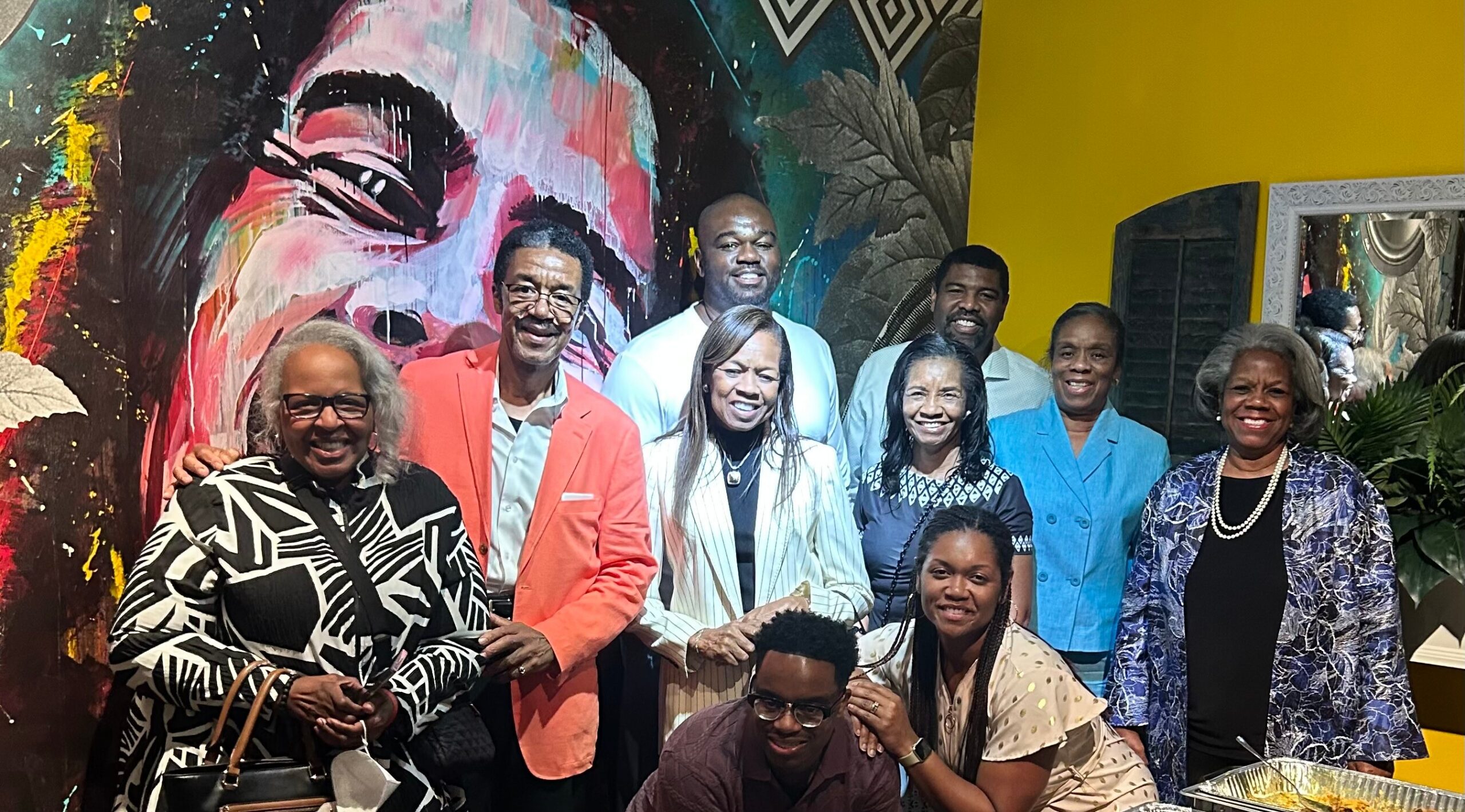 Group of diverse adults smiling for a group photo in front of a vivid mural; colorful clothing and a yellow wall are visible on the right.
