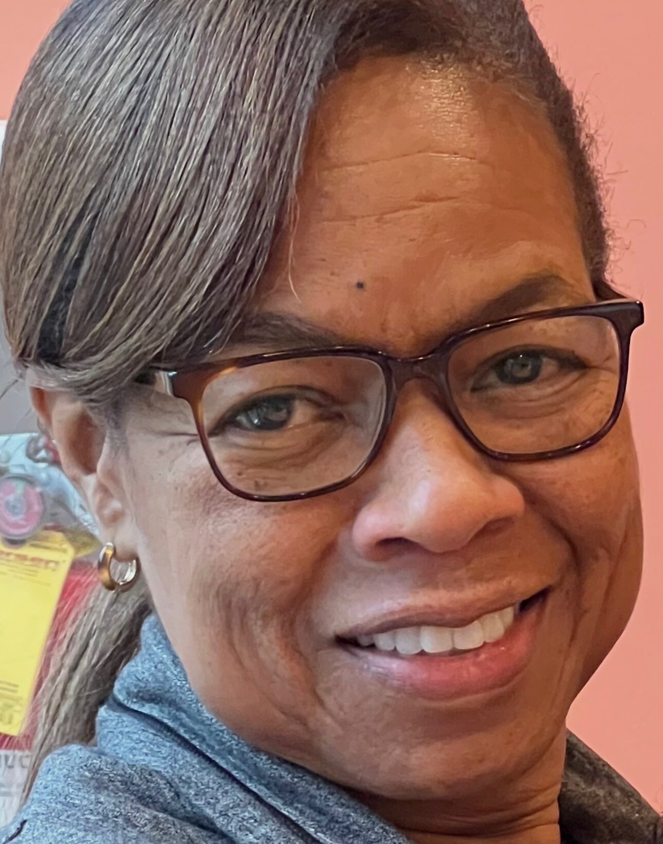 Close-up portrait of a smiling woman with dark glasses, gray streaked hair, and hoop earrings against a pink background.