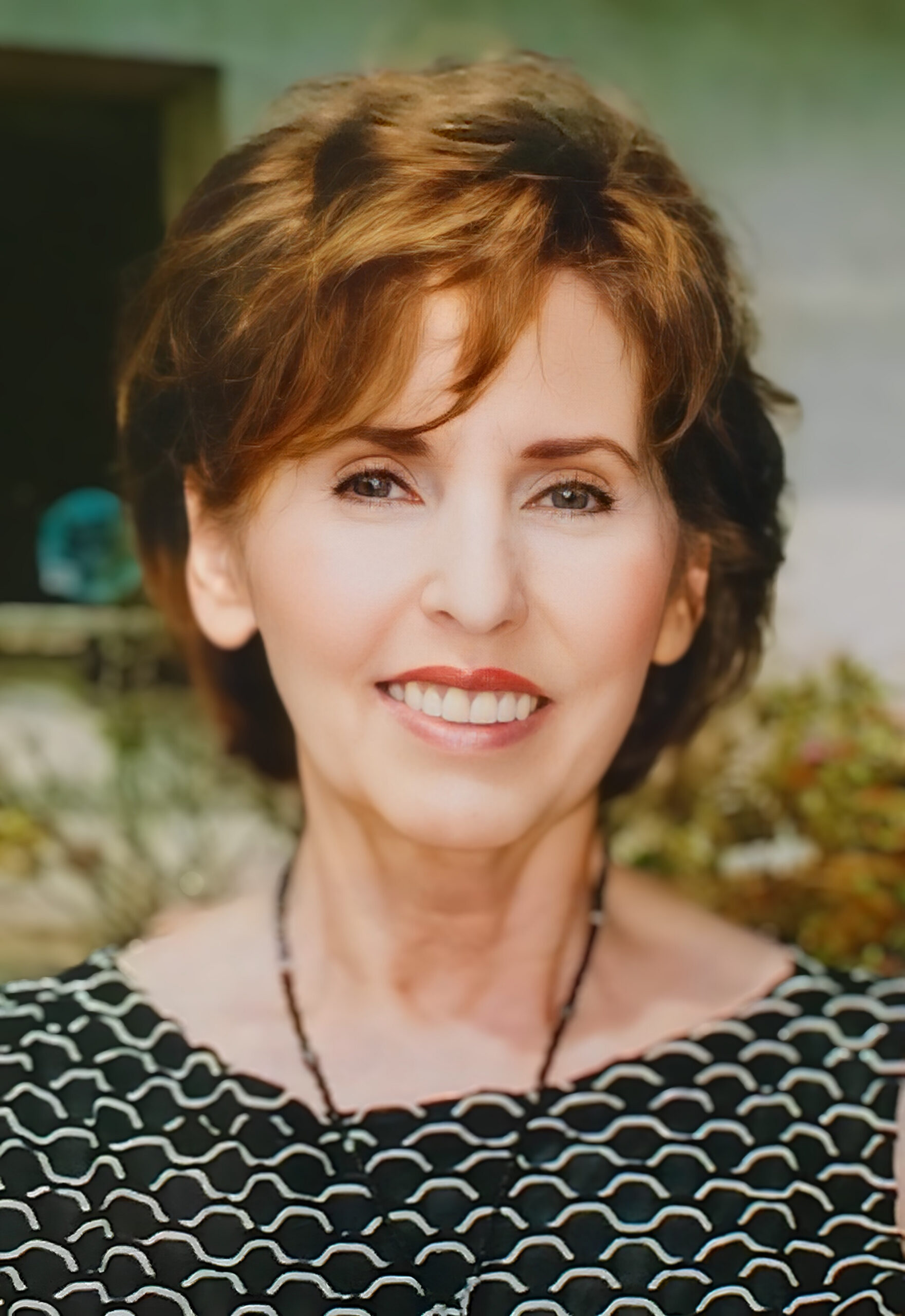 Close-up of a smiling woman with short brown hair, wearing a black-and-white wavy-patterned top, outdoors