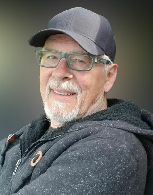 Smiling portrait of a man with a gray beard wearing a dark baseball cap and green glasses, looking at the camera.