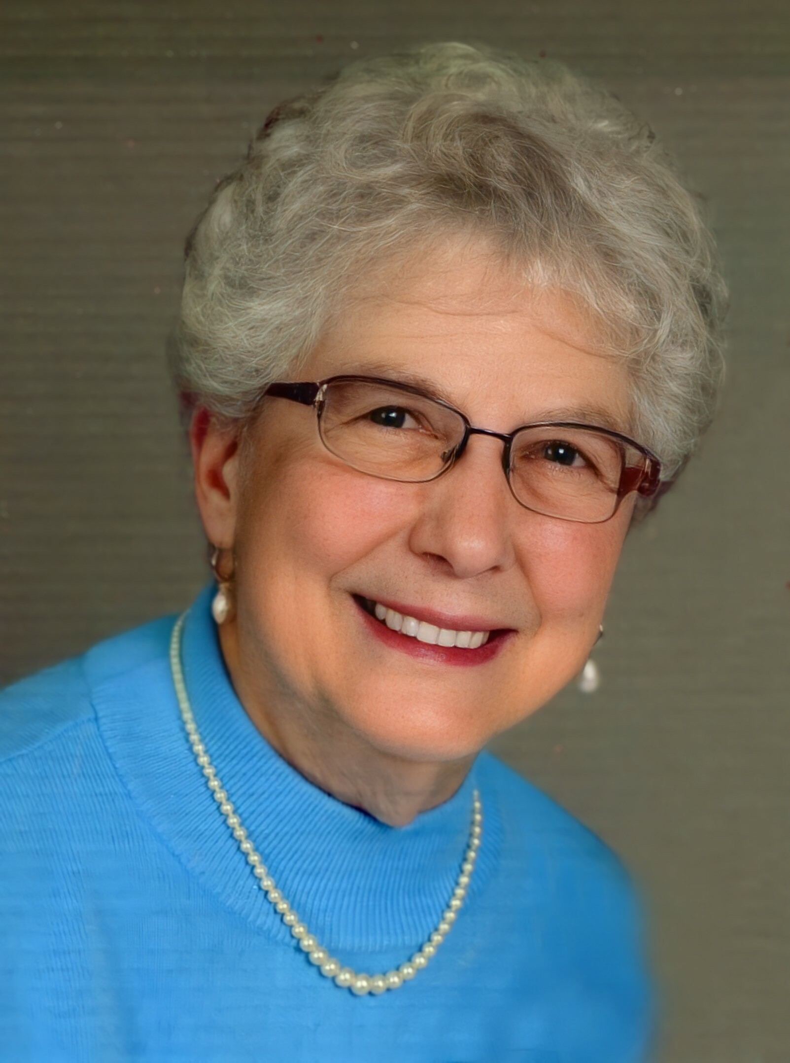 Smiling older woman with short gray hair wearing glasses, a blue shirt, and a pearl necklace.