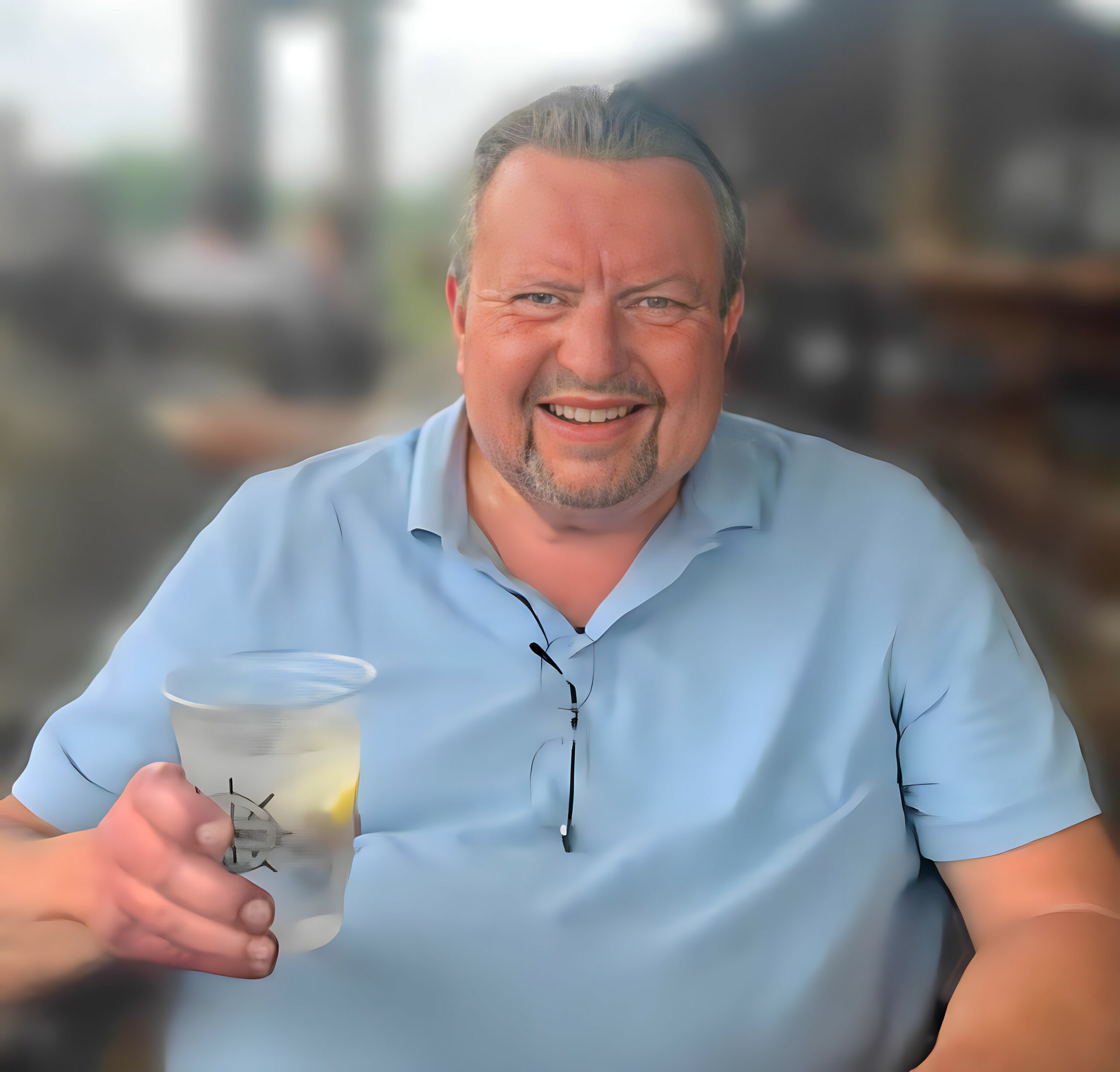 Smiling man in a light blue shirt, holding a baby bottle toward the camera in focus foreground with a blurred background