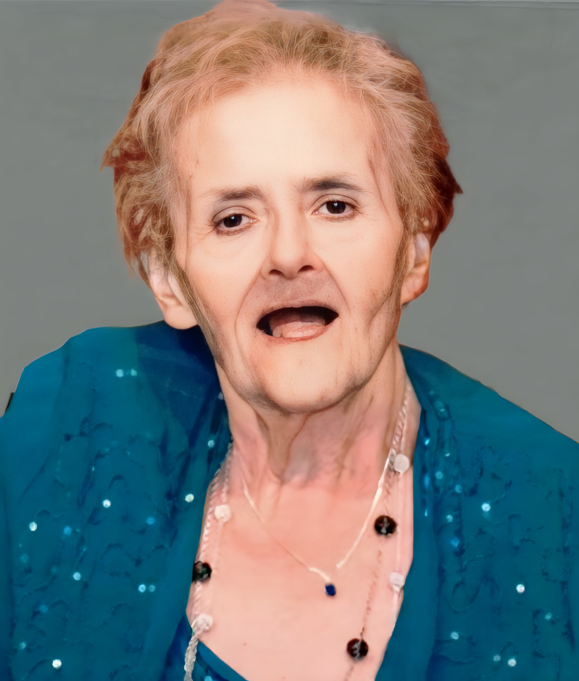 Close-up of a woman with short blond hair, mouth open as if singing, wearing a teal blouse.