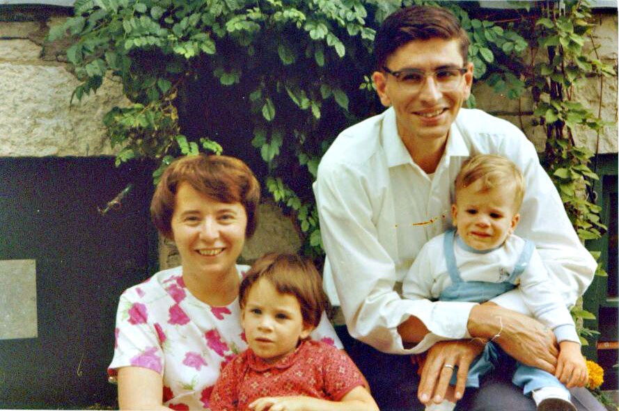 Family of four posing for a photo outdoors, mother with toddler on her lap and father holding a baby son in blue overalls, greenery behind them.
