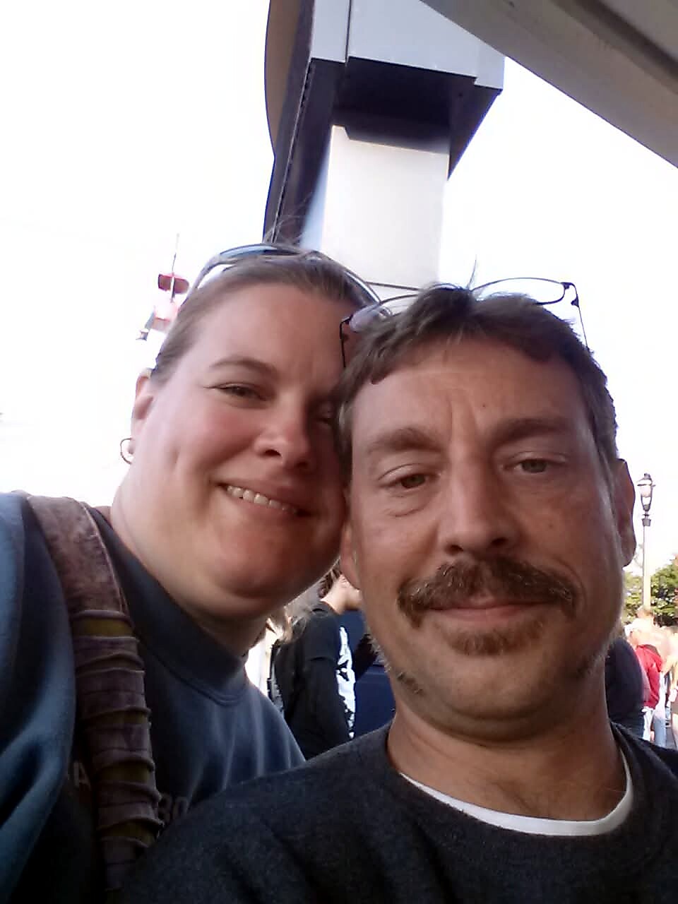 Two people smiling for a selfie outdoors, woman on the left wearing glasses on her head and a backpack strap visible, man on the right with a mustache.