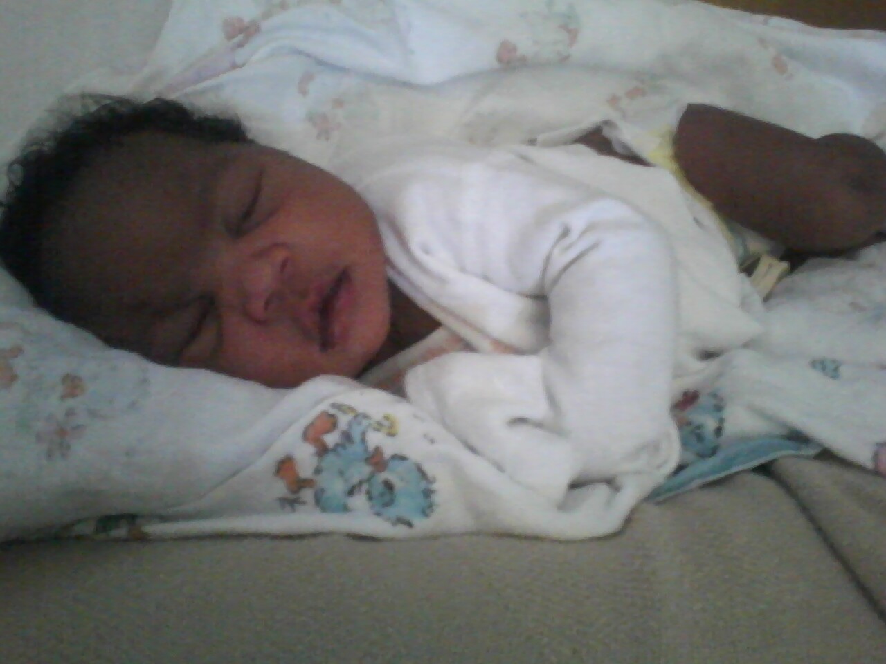 Newborn baby sleeping on a soft surface, wrapped in a white blanket with small colorful patterns.