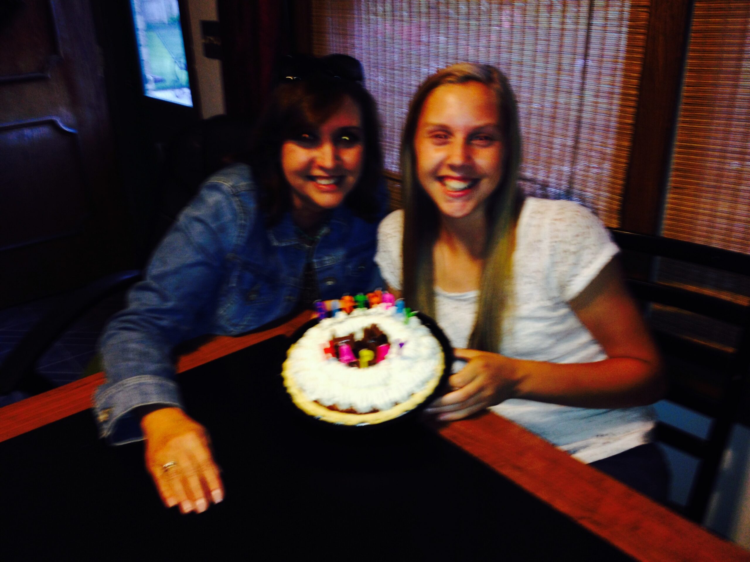Two smiling people sit at a table with a white frosted cake decorated with colorful candies in front of them.