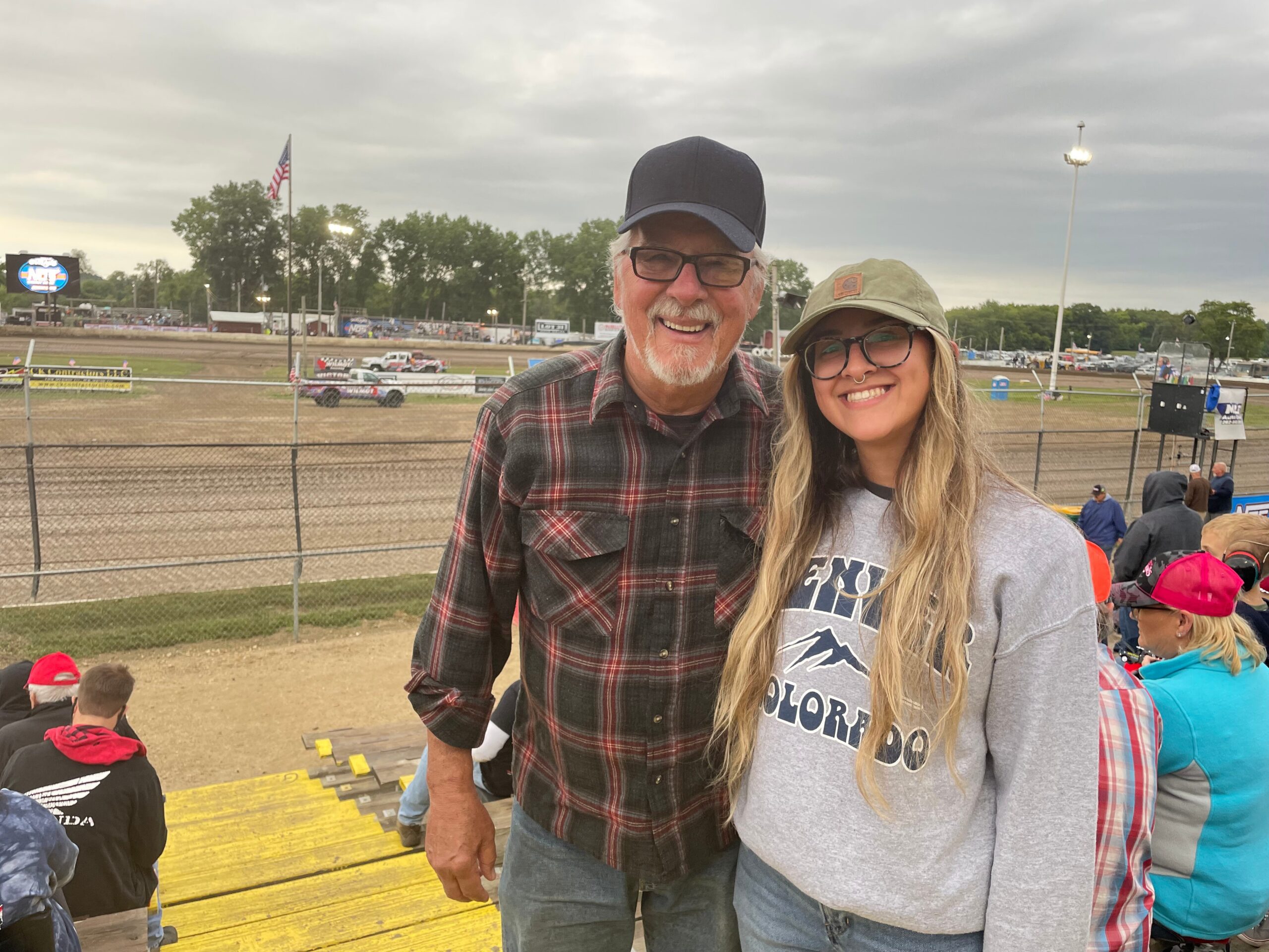 Two smiling people at a dirt racetrack: a man in a black cap and plaid shirt, and a woman in a gray hoodie, with bleachers behind them.