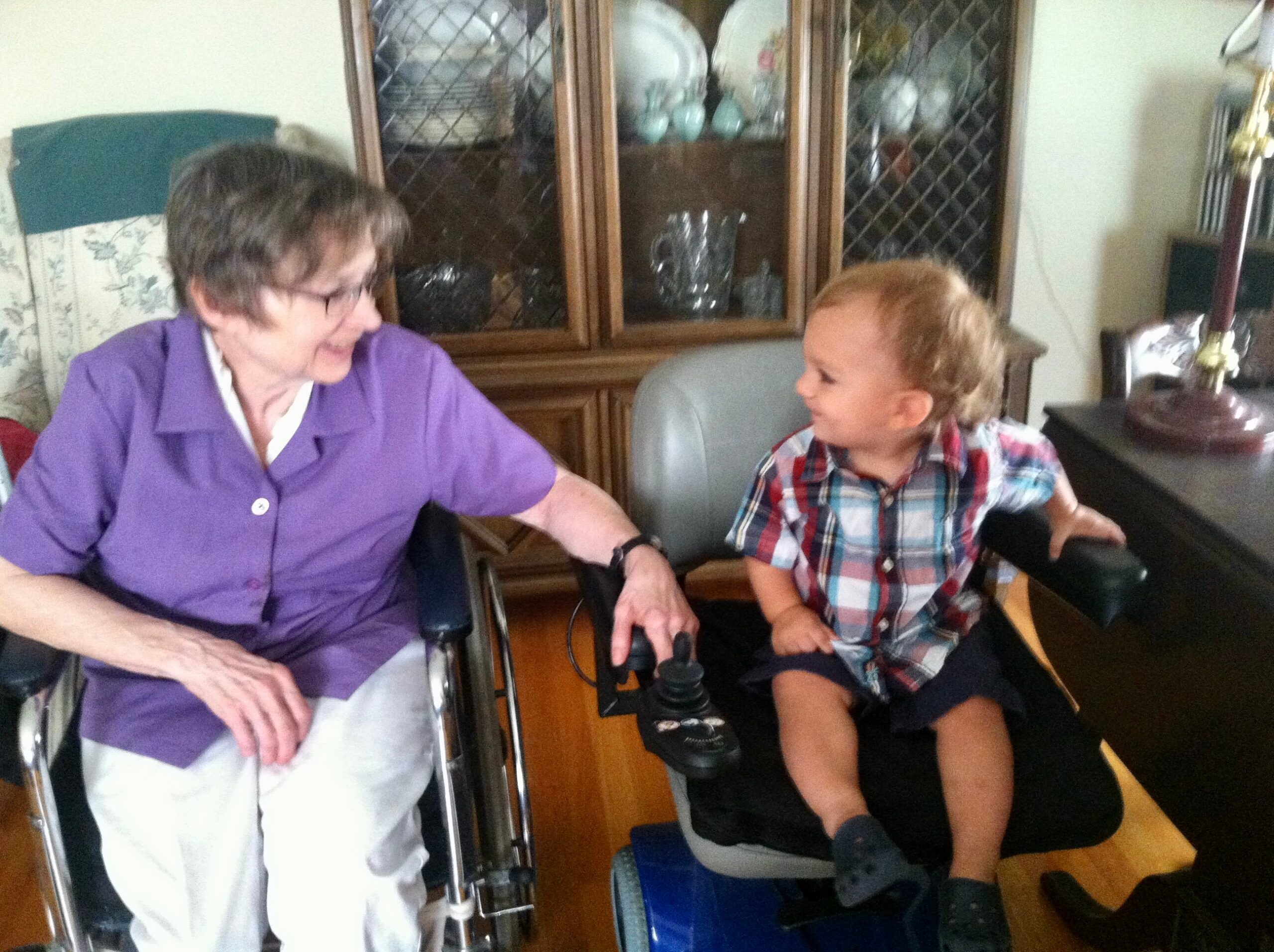 Older woman in a purple blouse sits in a wheelchair beside a smiling boy in a plaid shirt in a living room.