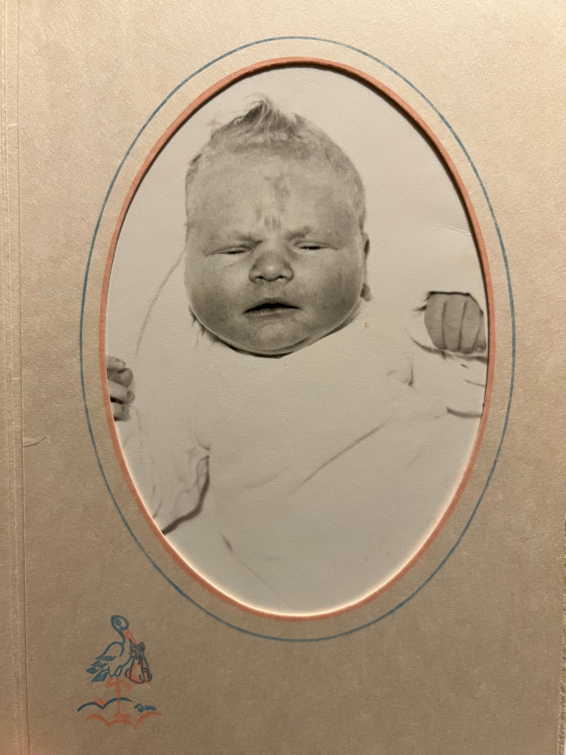 Vintage baby portrait in an oval frame on a beige textured page, with a small blue/red decorative illustration in the lower-left corner.