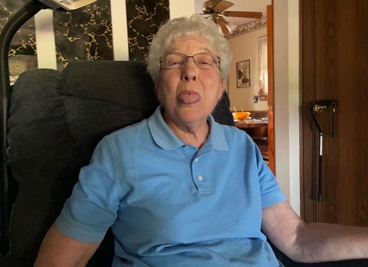 Older woman with short gray hair and glasses in a blue polo shirt, sticking out her tongue playfully in a home setting.