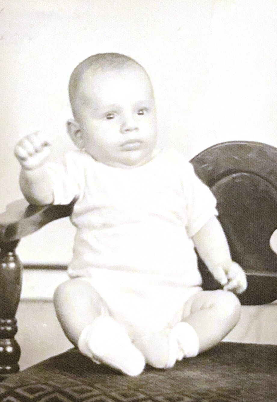 Infant seated on a rug, reaching with his right arm toward the camera.