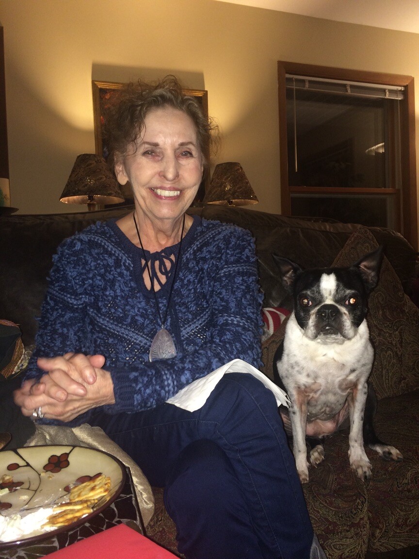 Smiling elderly woman in a blue sweater sits on a couch beside a small black-and-white dog with a plate of food visible in the foreground.