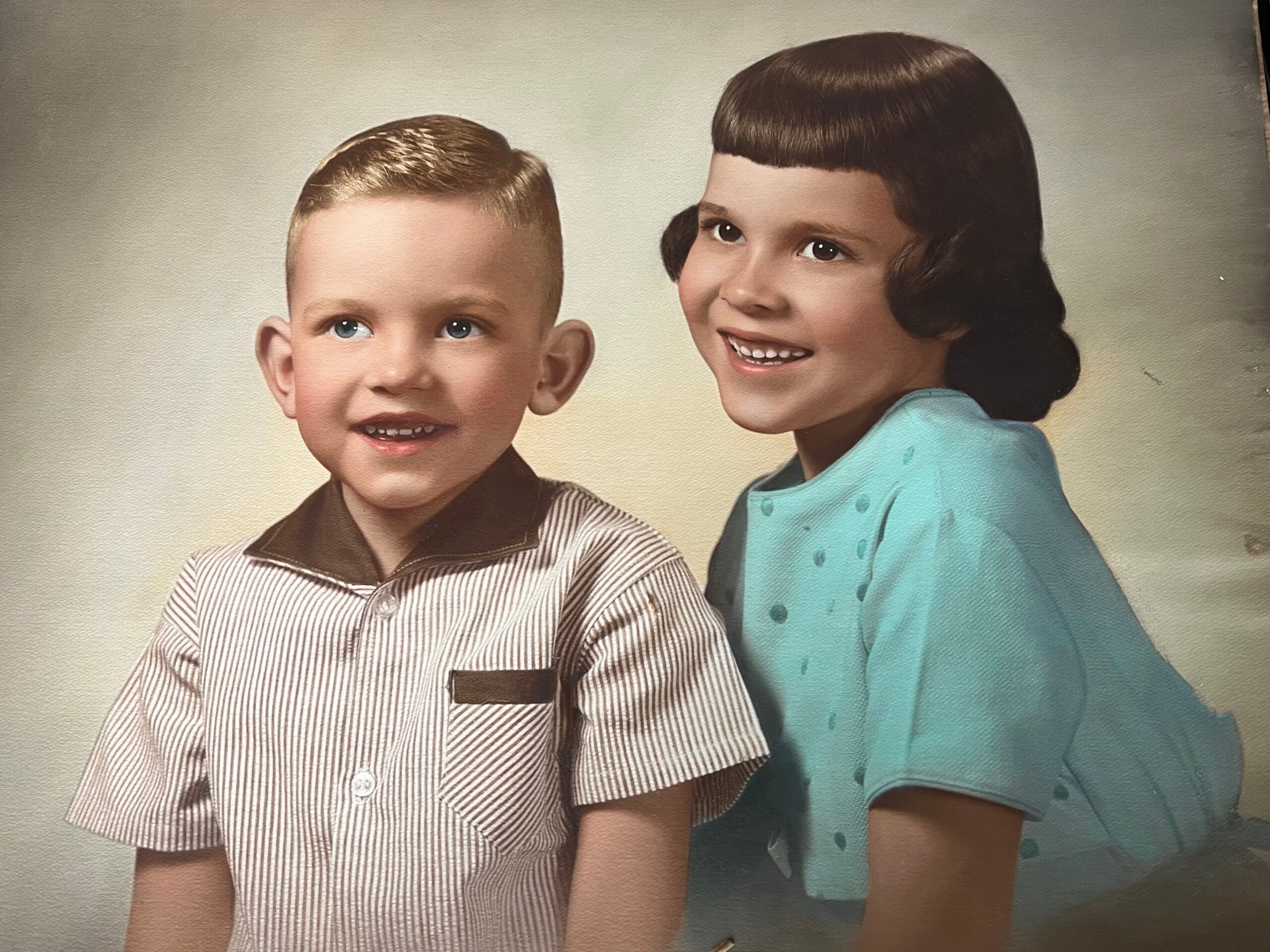 Two smiling children pose together in a studio portrait; a boy in a striped shirt and a girl in a teal dress side by side.