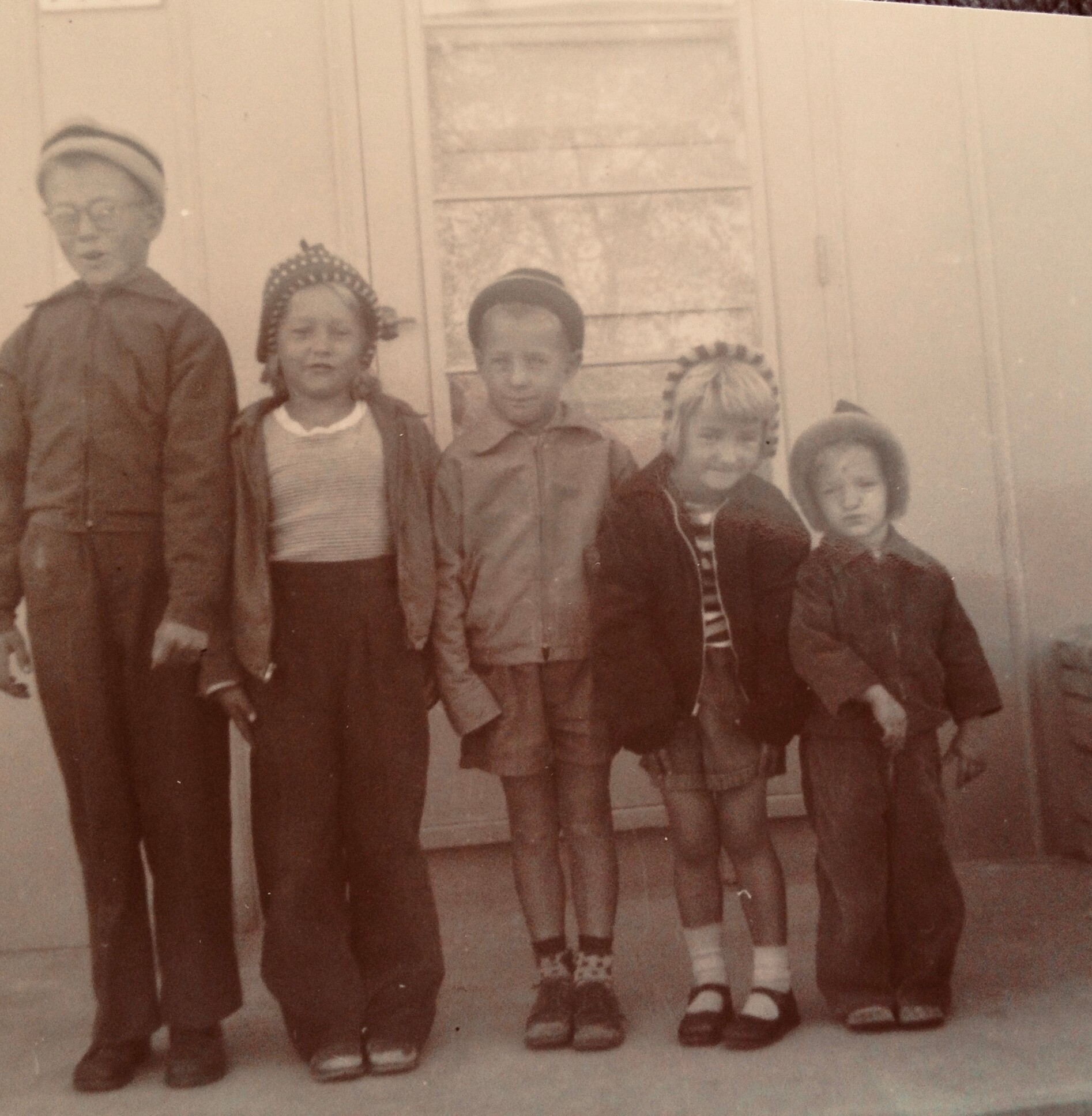 Five children stand in a row outside a doorway, wearing vintage coats and hats in a sepia-toned photo.