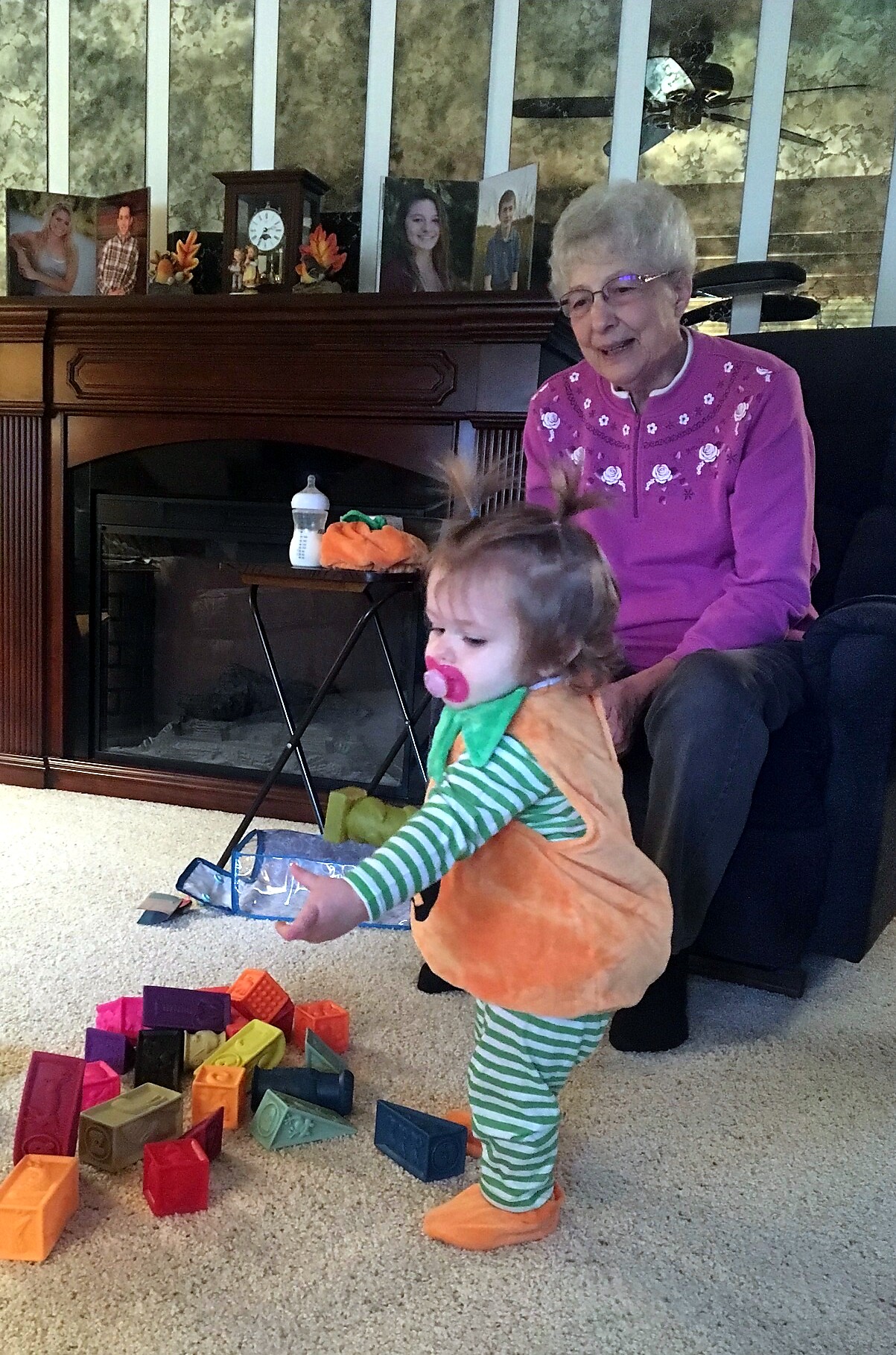 Grandmother sits on a chair watching a toddler in an animal costume stack colorful foam blocks on a carpeted floor near a fireplace with family photos on the mantel.