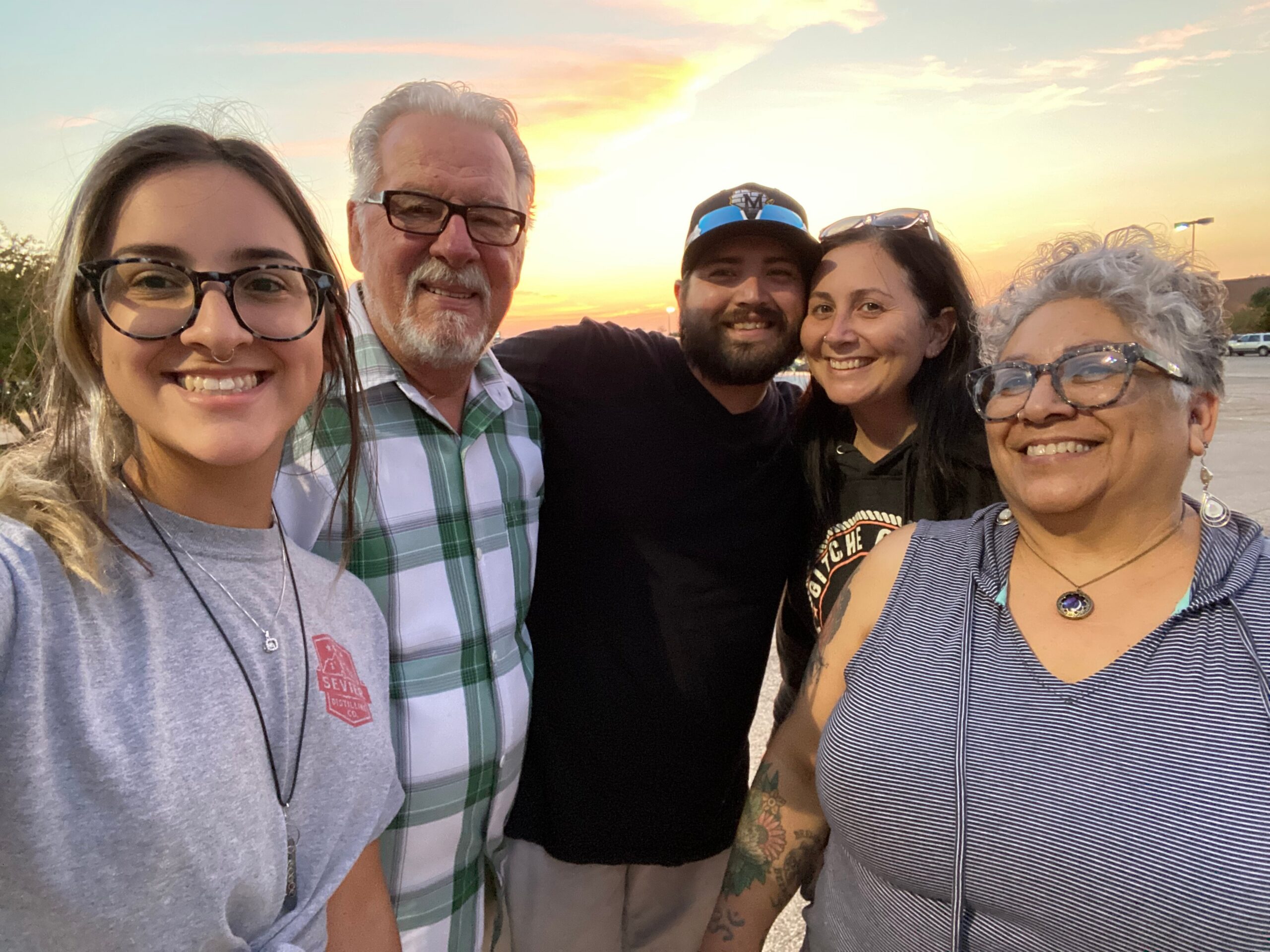 Group of five people smiling for a selfie at sunset in an outdoor parking lot. Sunlit sky with warm colors in the background, casual clothing and sunglasses on some, close together for the photo.