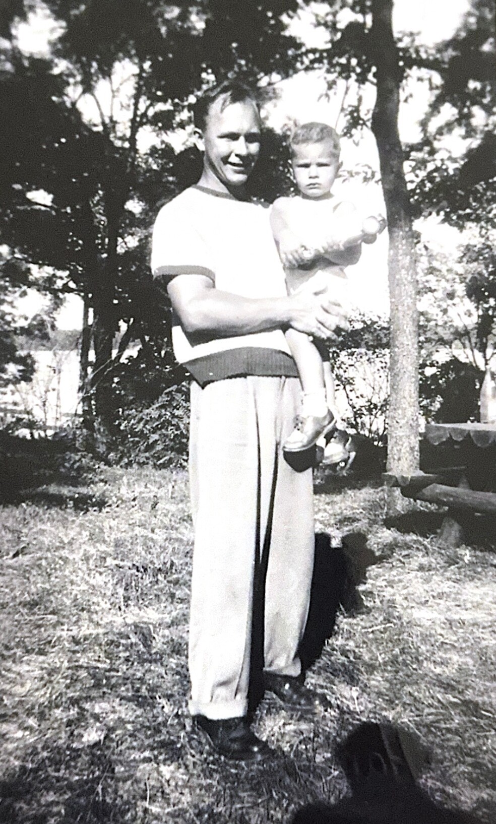 Father in a light shirt holds a young child outdoors in a grassy, tree-filled yard.