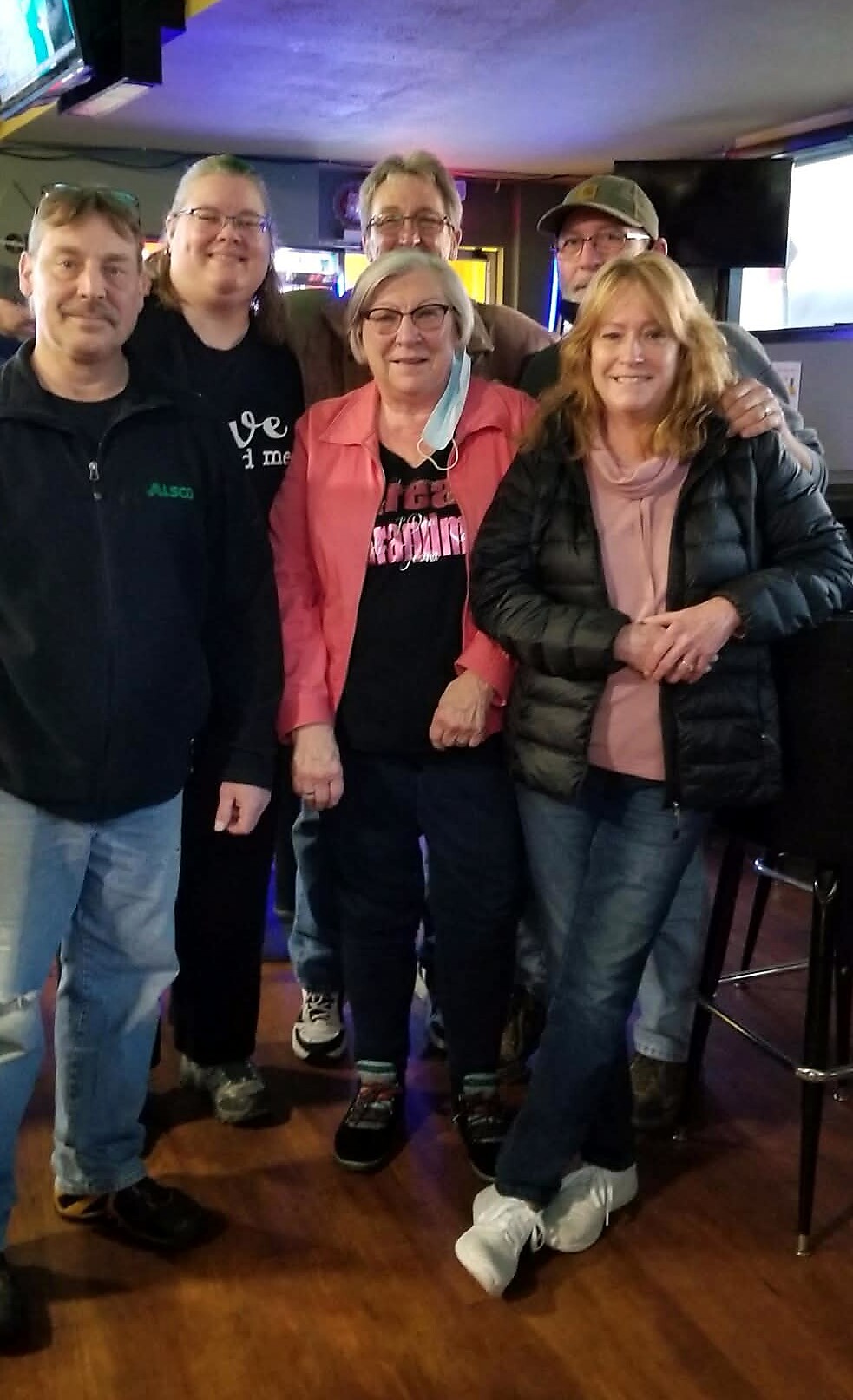 Five adults posing together in a dim bar, smiling at the camera; center person wears a pink jacket, others in casual jackets and jeans.