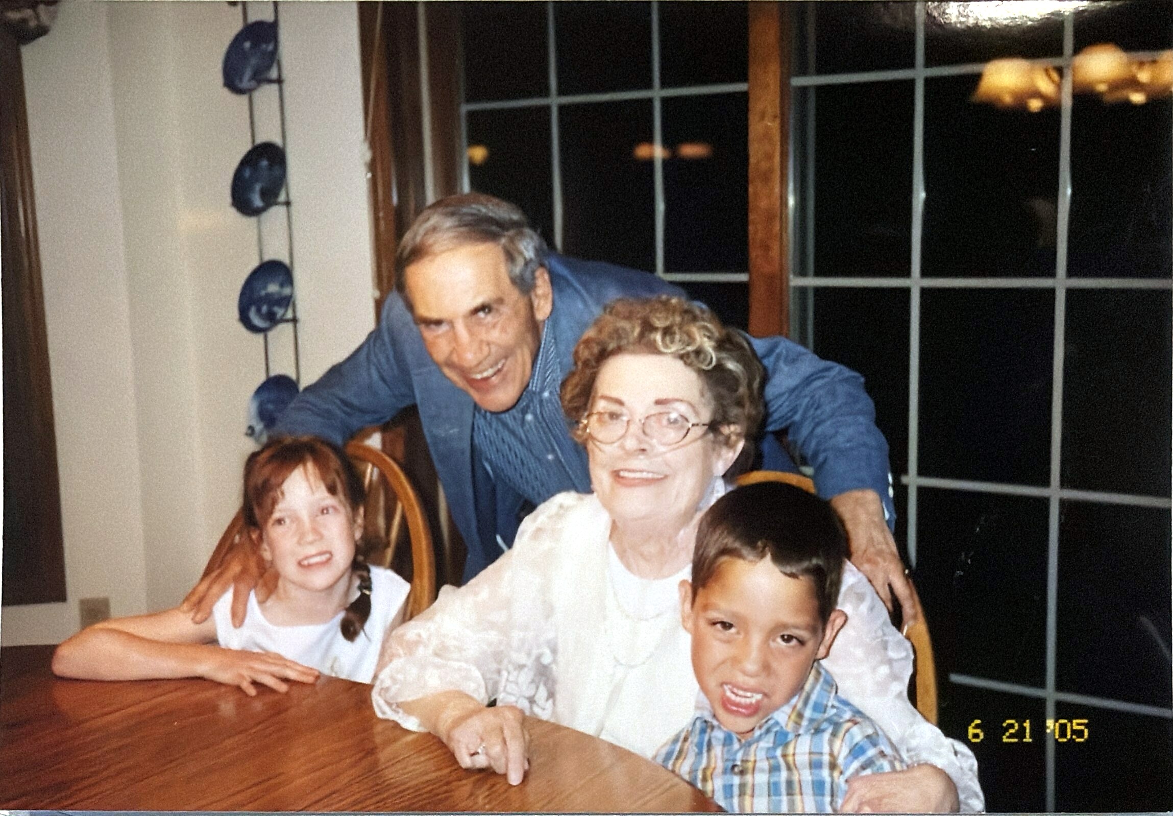 An older man leaning over a group of smiling children seated around a wooden table indoors, posing for a family photo.