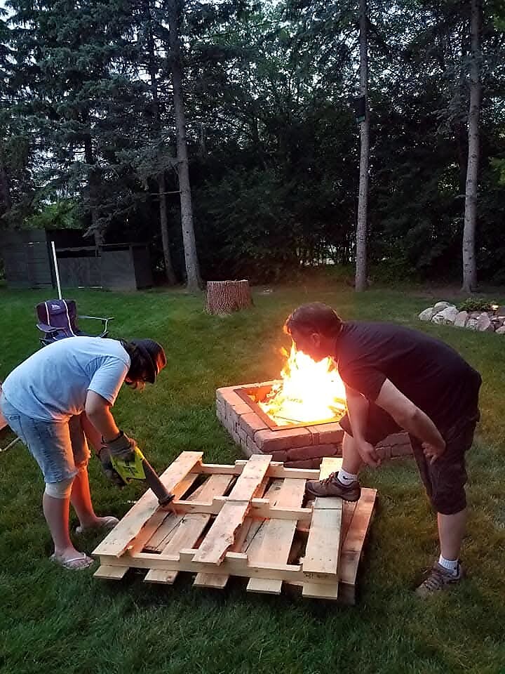 Two people hammering apart a wooden pallet beside a lit brick fire pit in a backyard.
