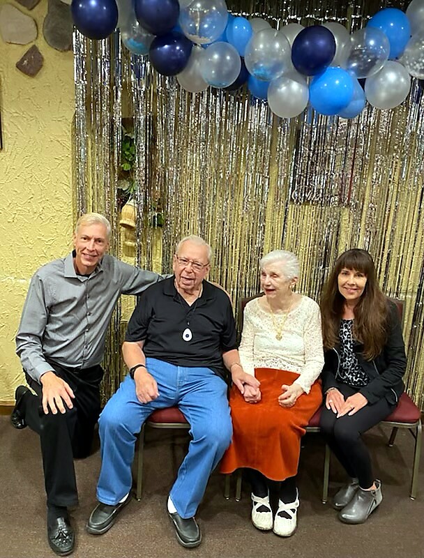 Four people sit side by side on chairs at a celebration, smiling, with blue and silver balloons and a metallic fringe backdrop above them.