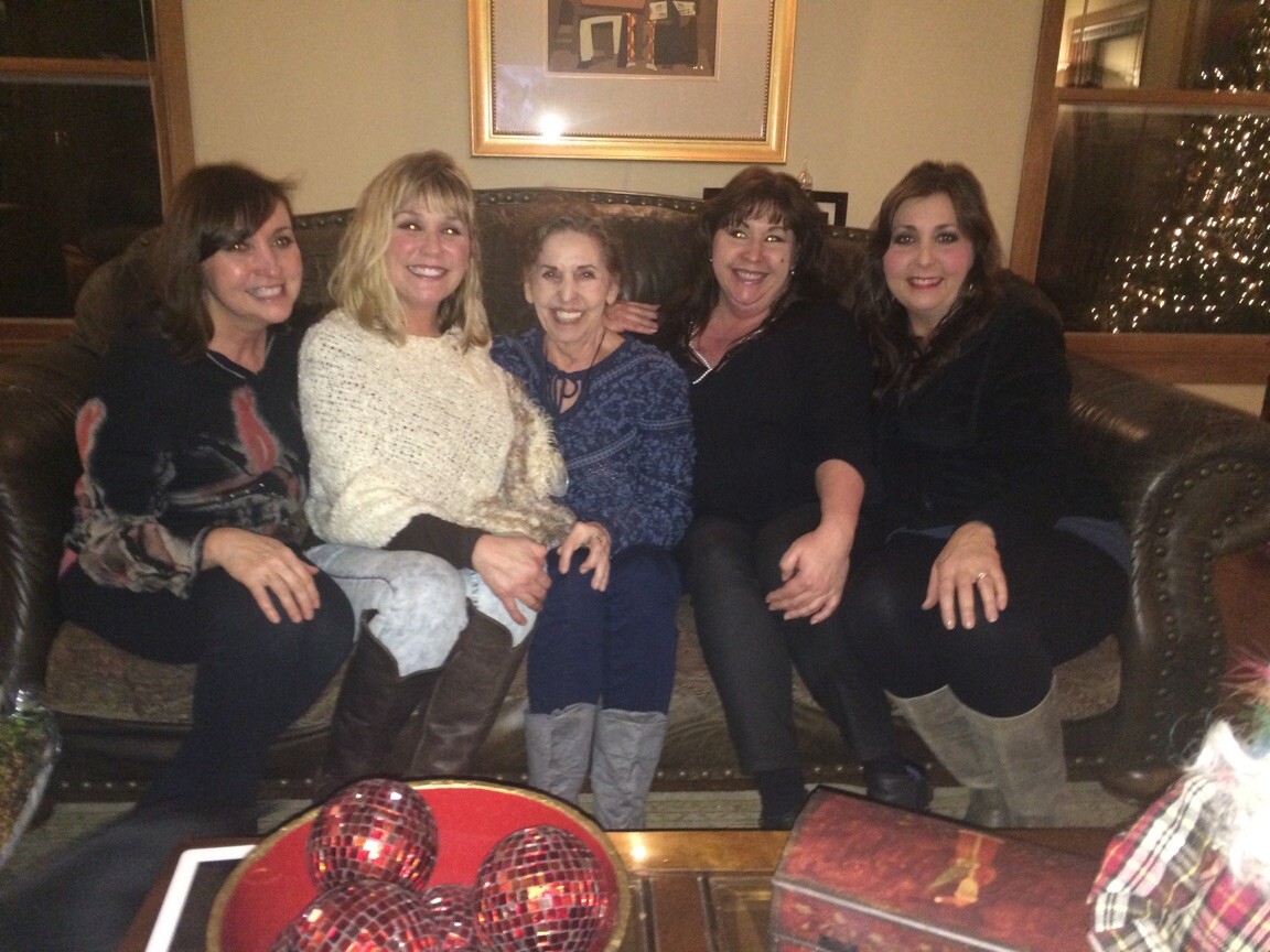 Five women sitting on a brown leather couch in a living room, smiling with a Christmas tree lit in the background.