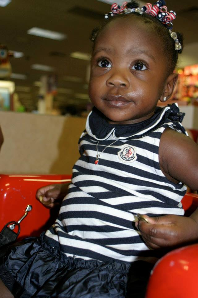Young girl wearing a navy-and-white striped sleeveless top with a bow headband, seated in a red toy vehicle indoors.