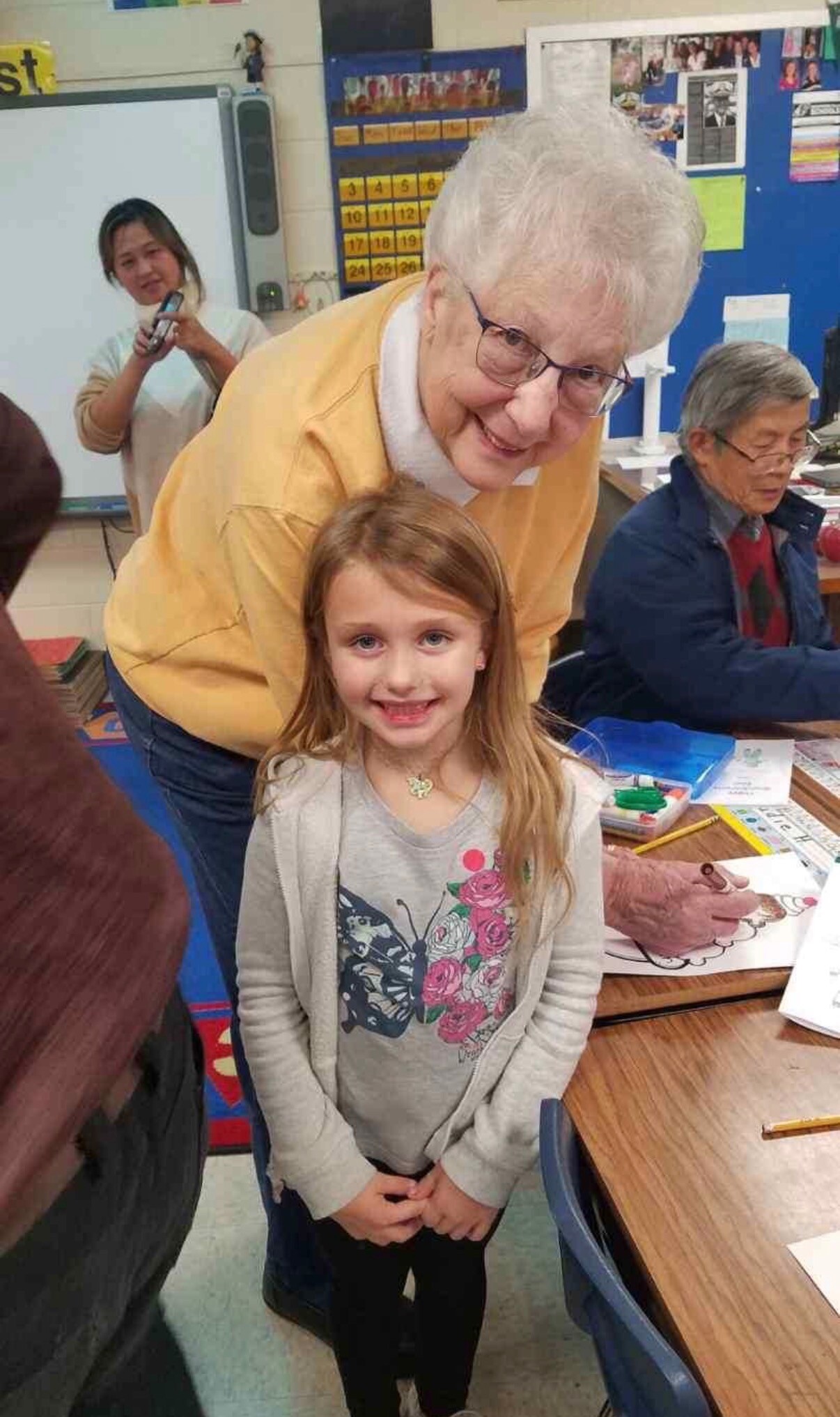 Grandmother leans toward a smiling young girl during a classroom drawing activity, with a teacher and other students in the background.