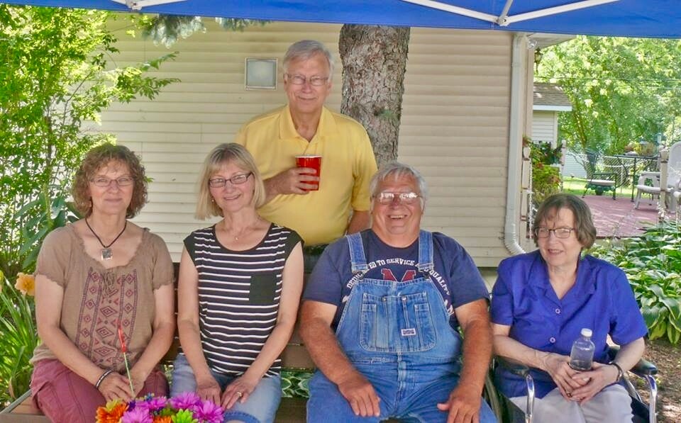 Group of five smiling adults posing under a blue canopy outdoors, with flowers in the foreground and a house behind them.