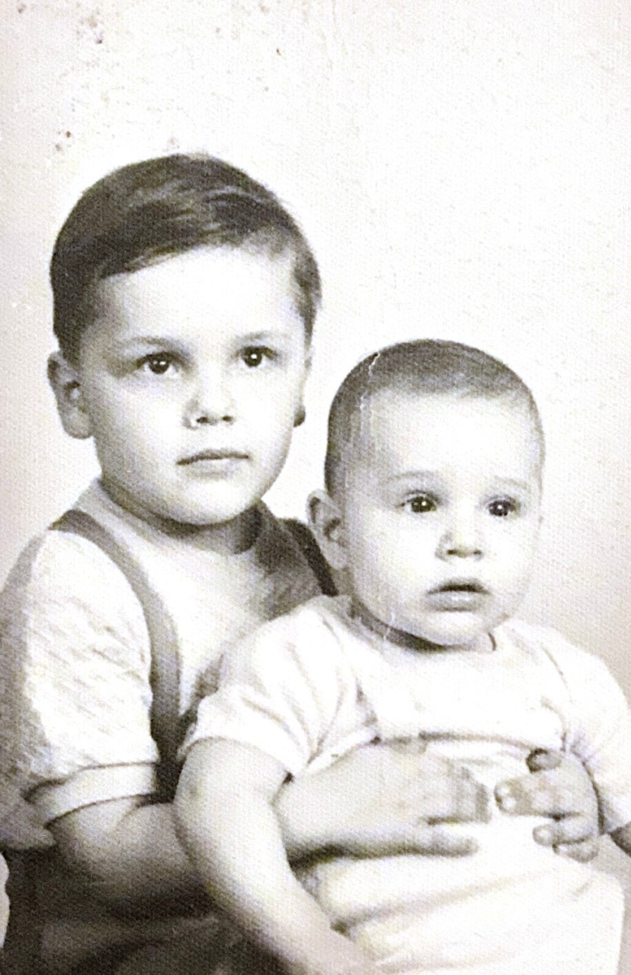 Two young siblings pose for a black-and-white portrait, the older boy holding the baby on his lap.