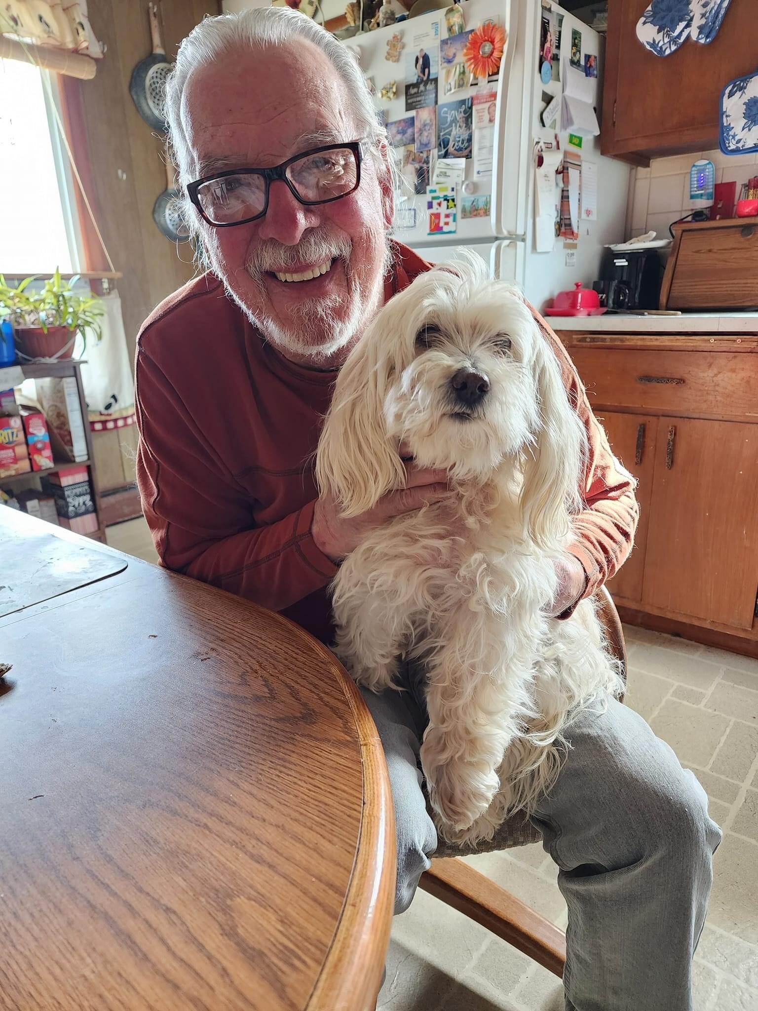 Smiling elderly man with glasses holding a fluffy white dog on his lap at a kitchen table in a cozy home setting.