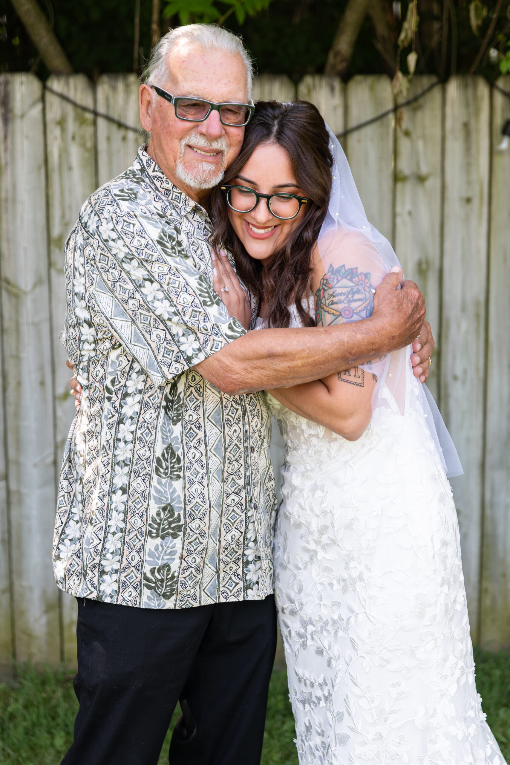 Older man in a tropical-print shirt hugs a smiling bride in a white lace wedding gown outdoors.