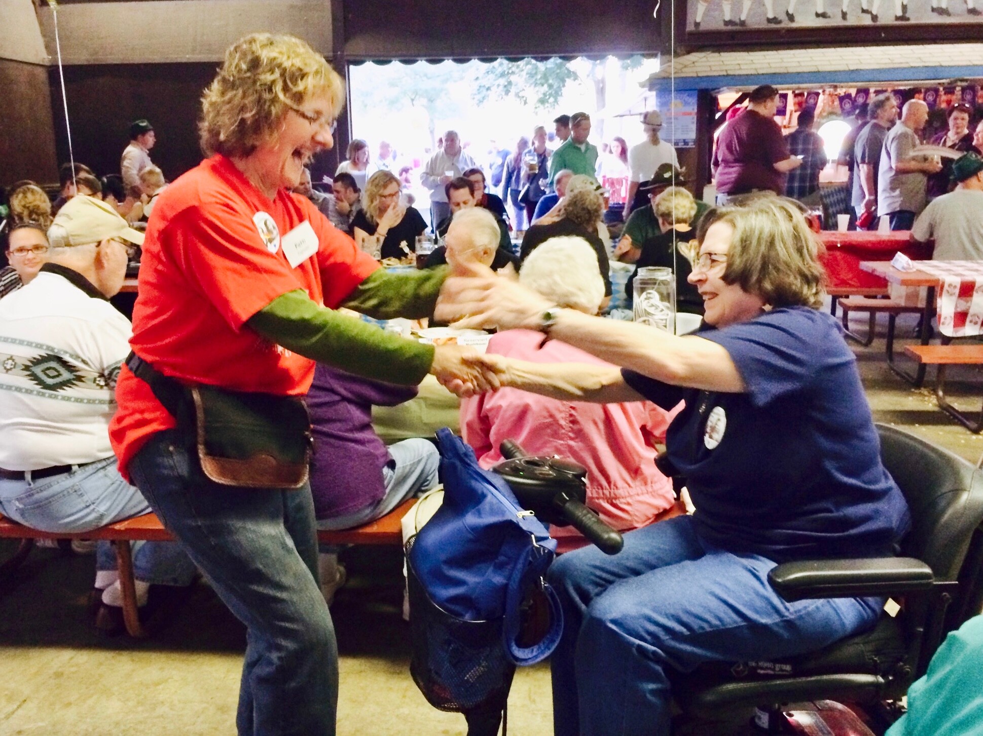 Two women shake hands at a busy indoor gathering; one stands in a red shirt, the other sits in a wheelchair at a community event.