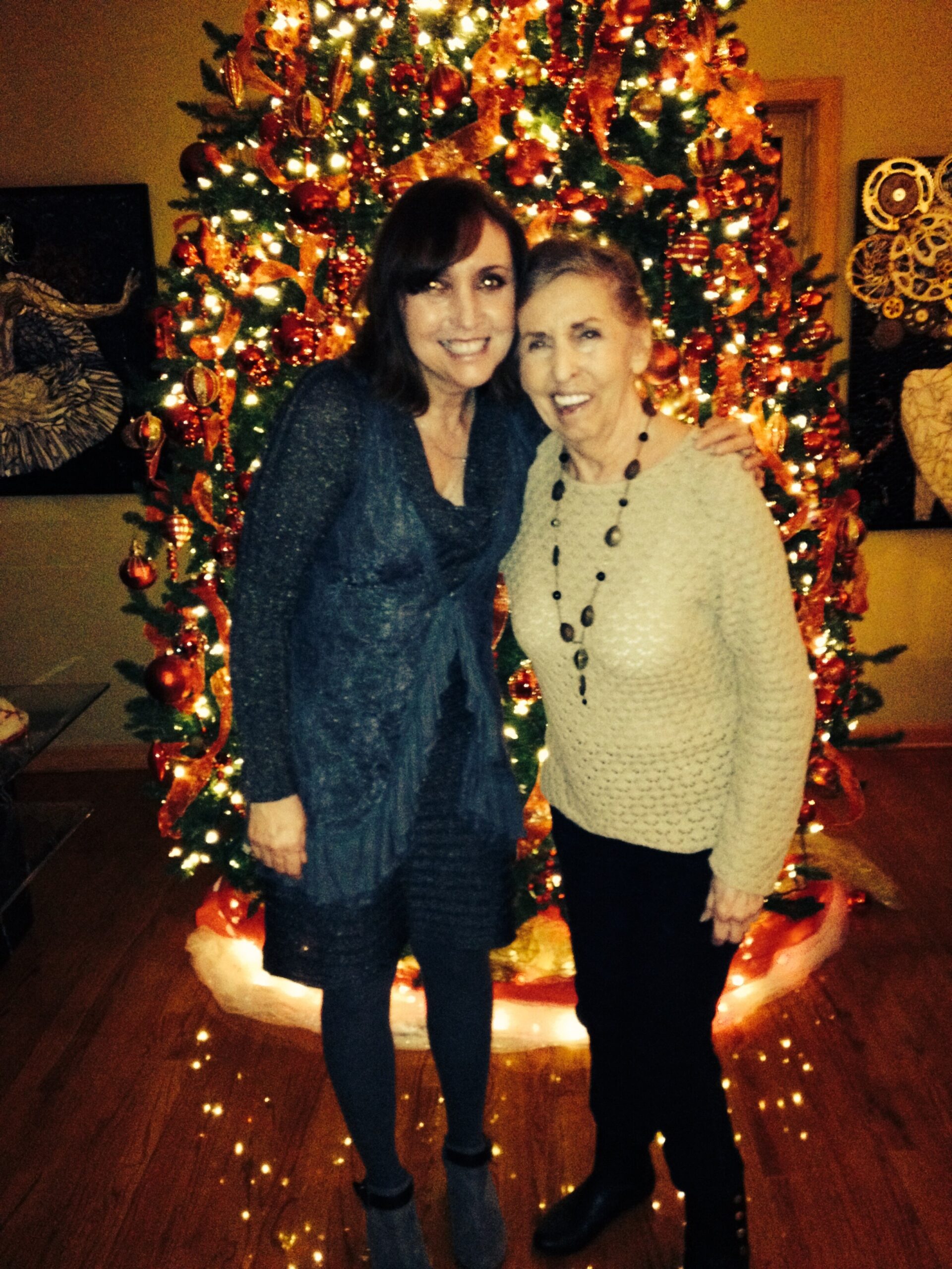 Two women pose arm in arm in front of a decorated Christmas tree with warm lights and red ornaments.