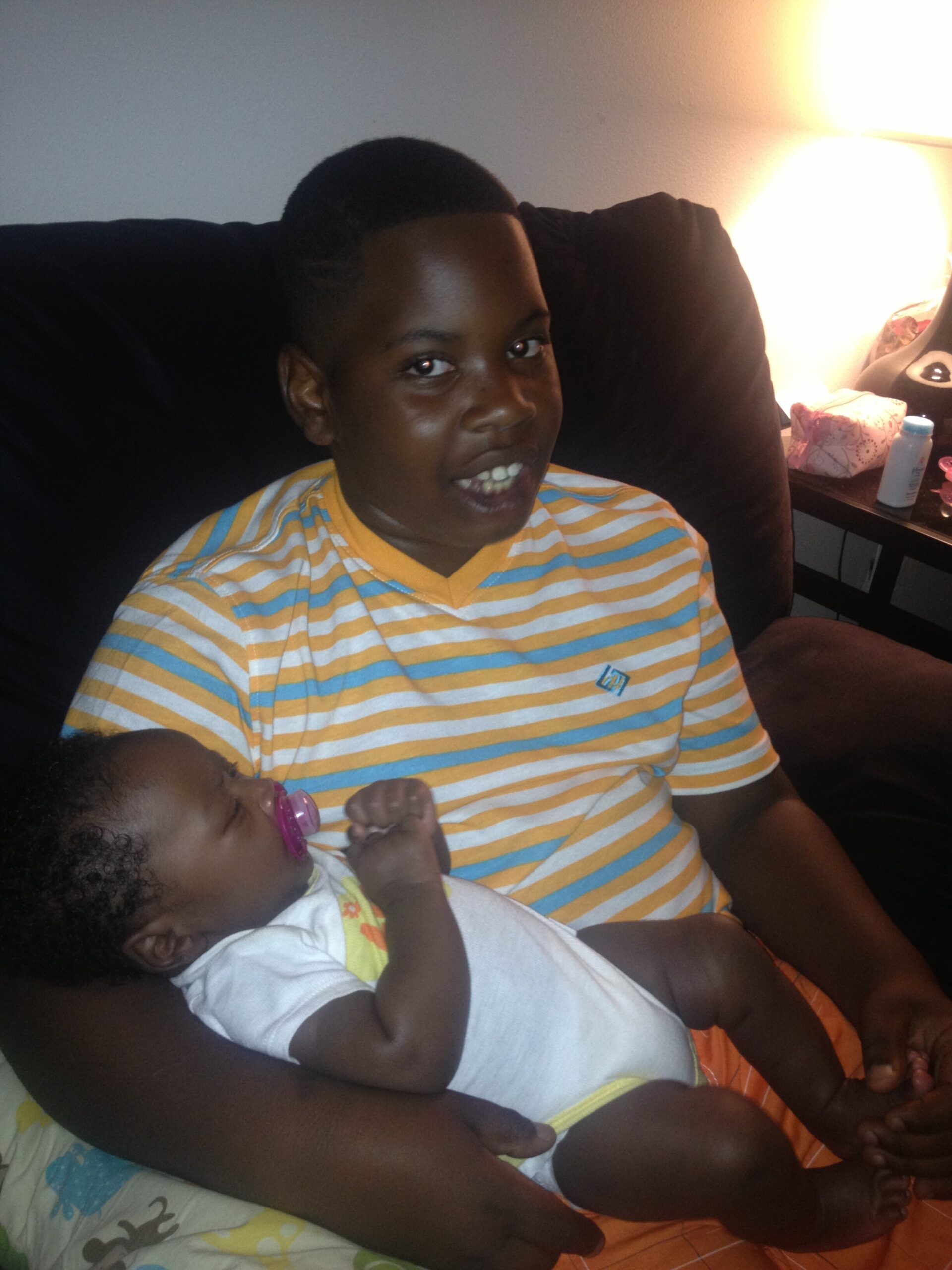 Two children sitting on a dark sofa: an older child in a yellow, blue, and white striped shirt smiles at the camera while holding a baby with a pink pacifier. A nightstand with a lamp and items sits in the background.