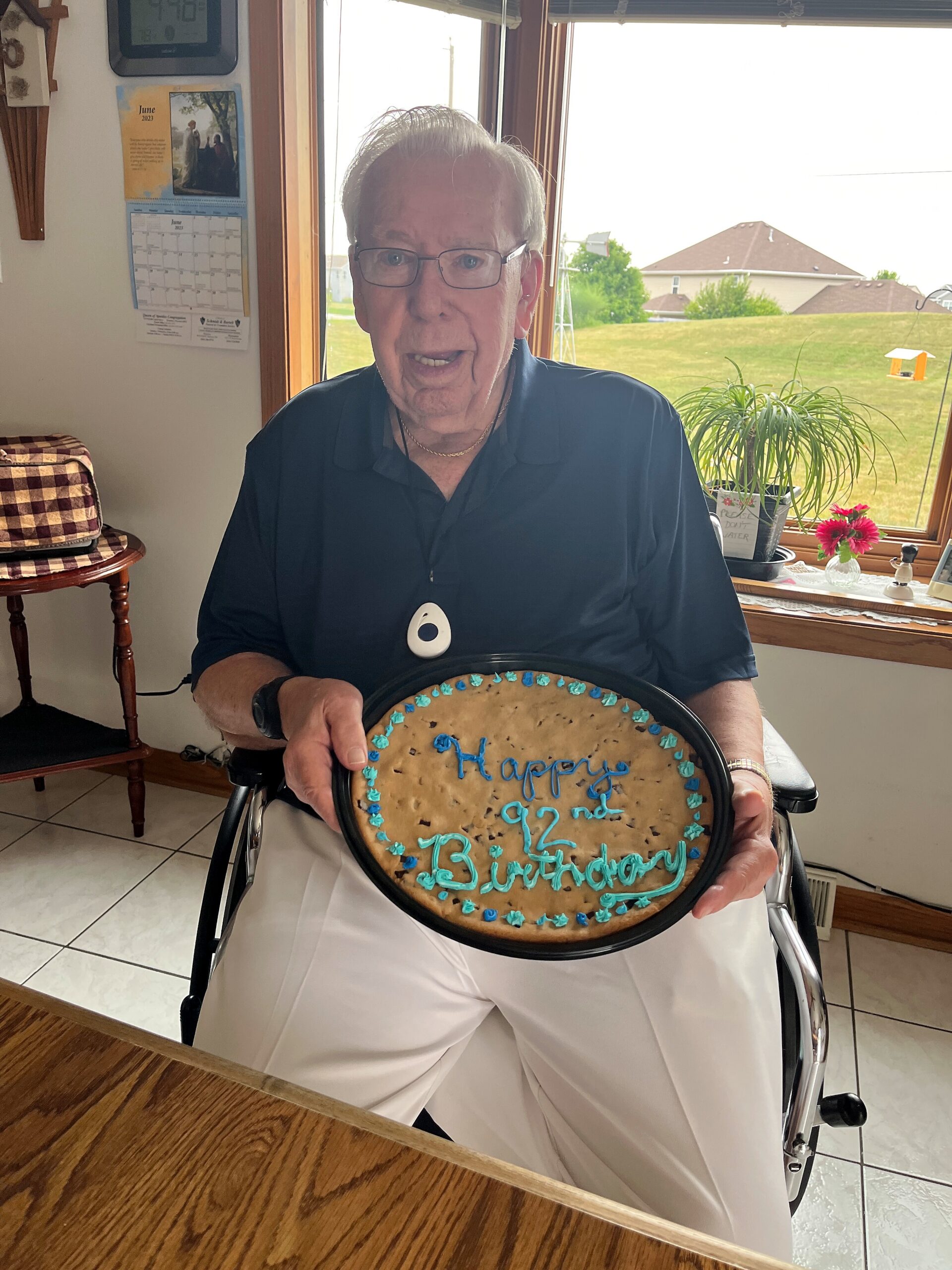 Elderly man in a wheelchair holding a large cake that reads 'Happy 92nd Birthday' with blue decorations in a sunny kitchen window setting.