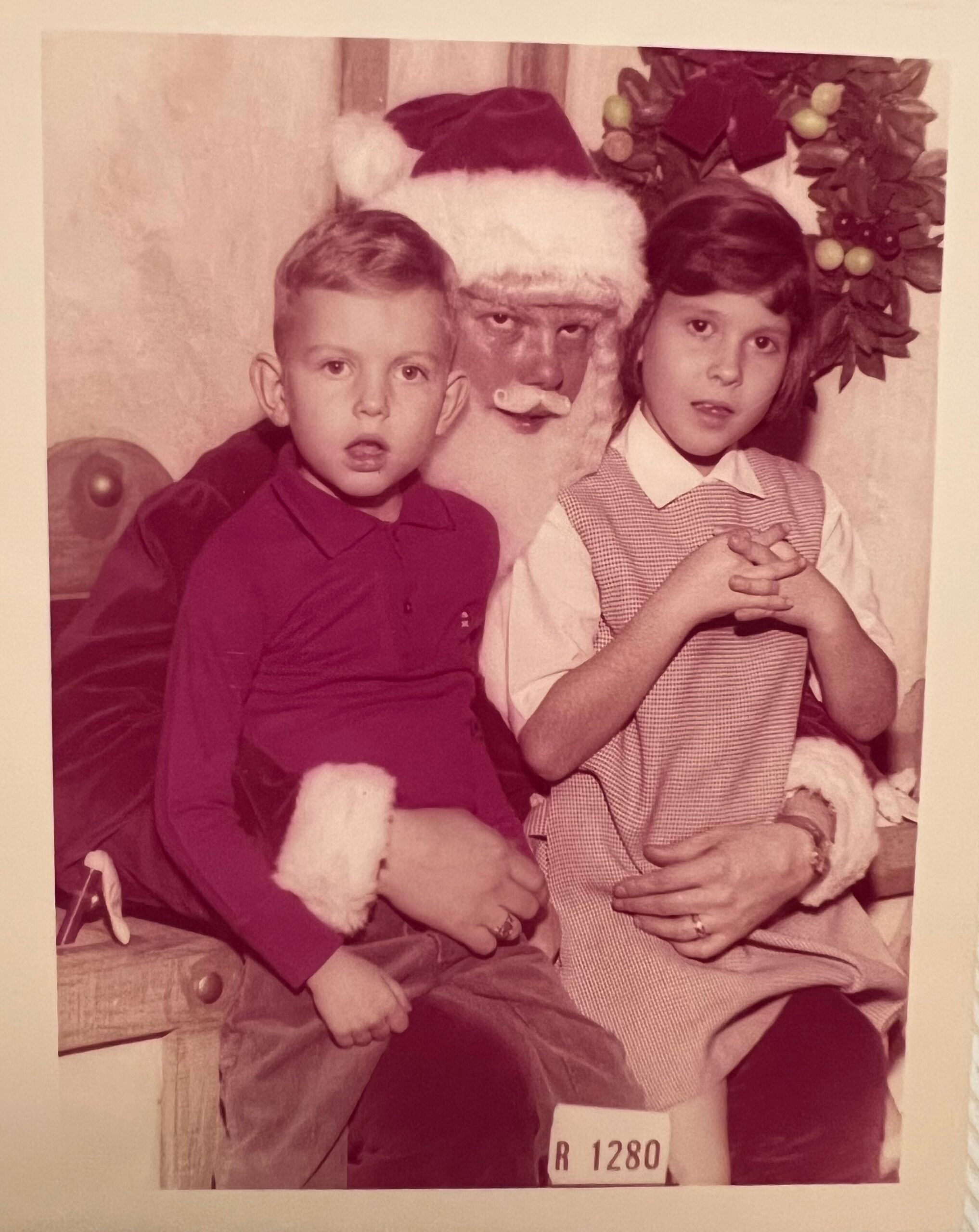 Two children sit with Santa Claus for a Christmas photo; the boy on the left wears a red shirt and the girl on the right wears a checkered dress, with Santa between them.