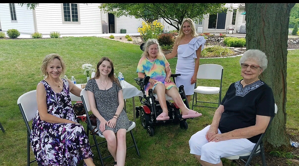 Five women sit and stand around a grassy yard, smiling for a photo near a table with water bottles; one woman is in a motorized wheelchair.